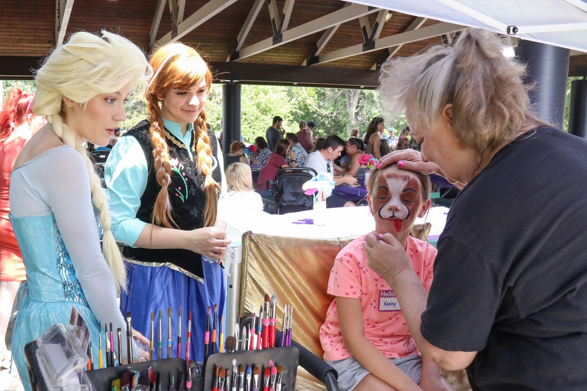 Face painter applying dog face to child, Elsa and Anna watch. Outdoor event, sunny.