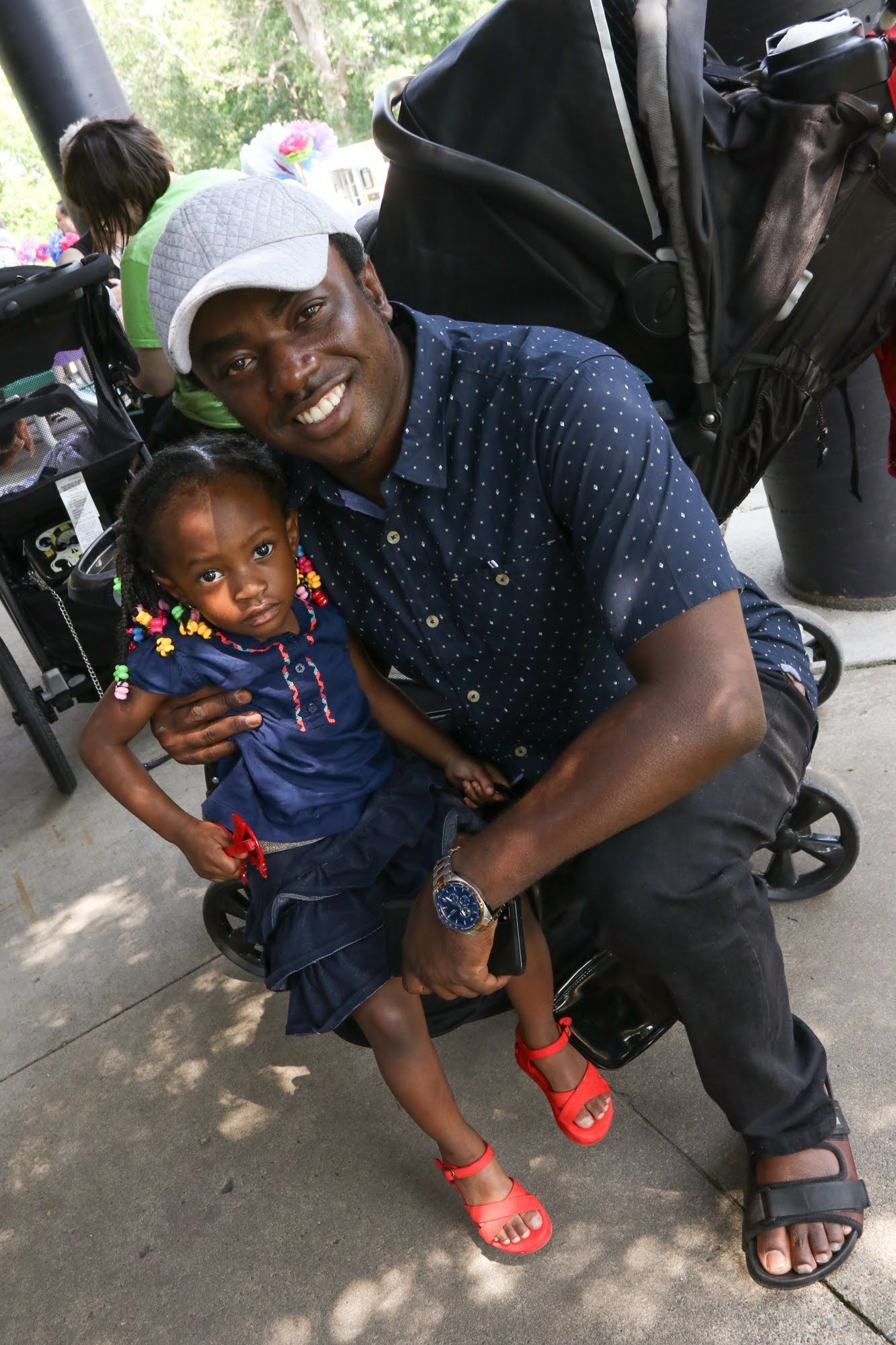 Man holds a child, smiling, by a stroller outdoors. The child wears a blue dress and red shoes.
