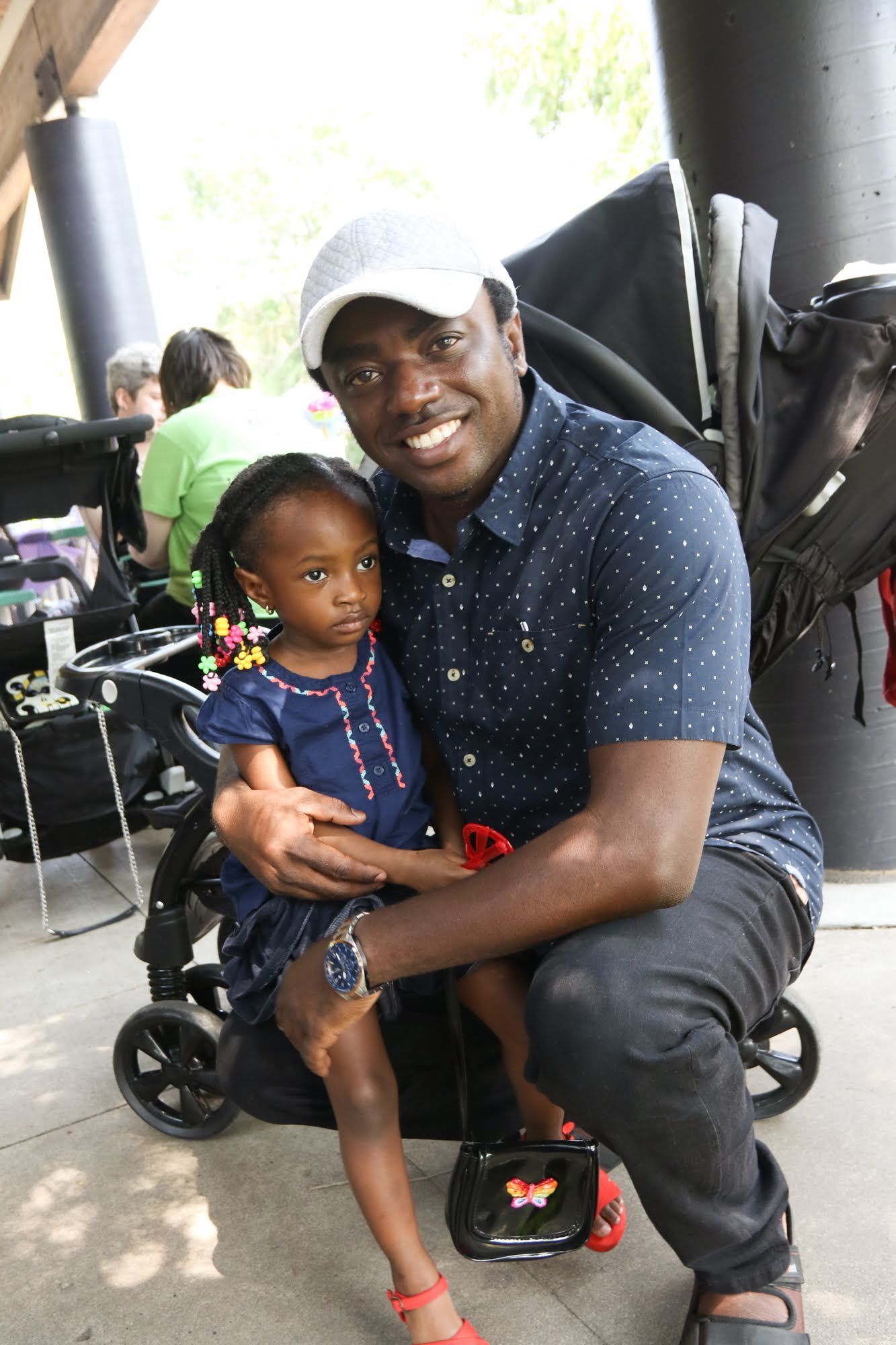 Man hugging a young child in front of a stroller; both smiling. Outdoors, blue shirt, gray hat.