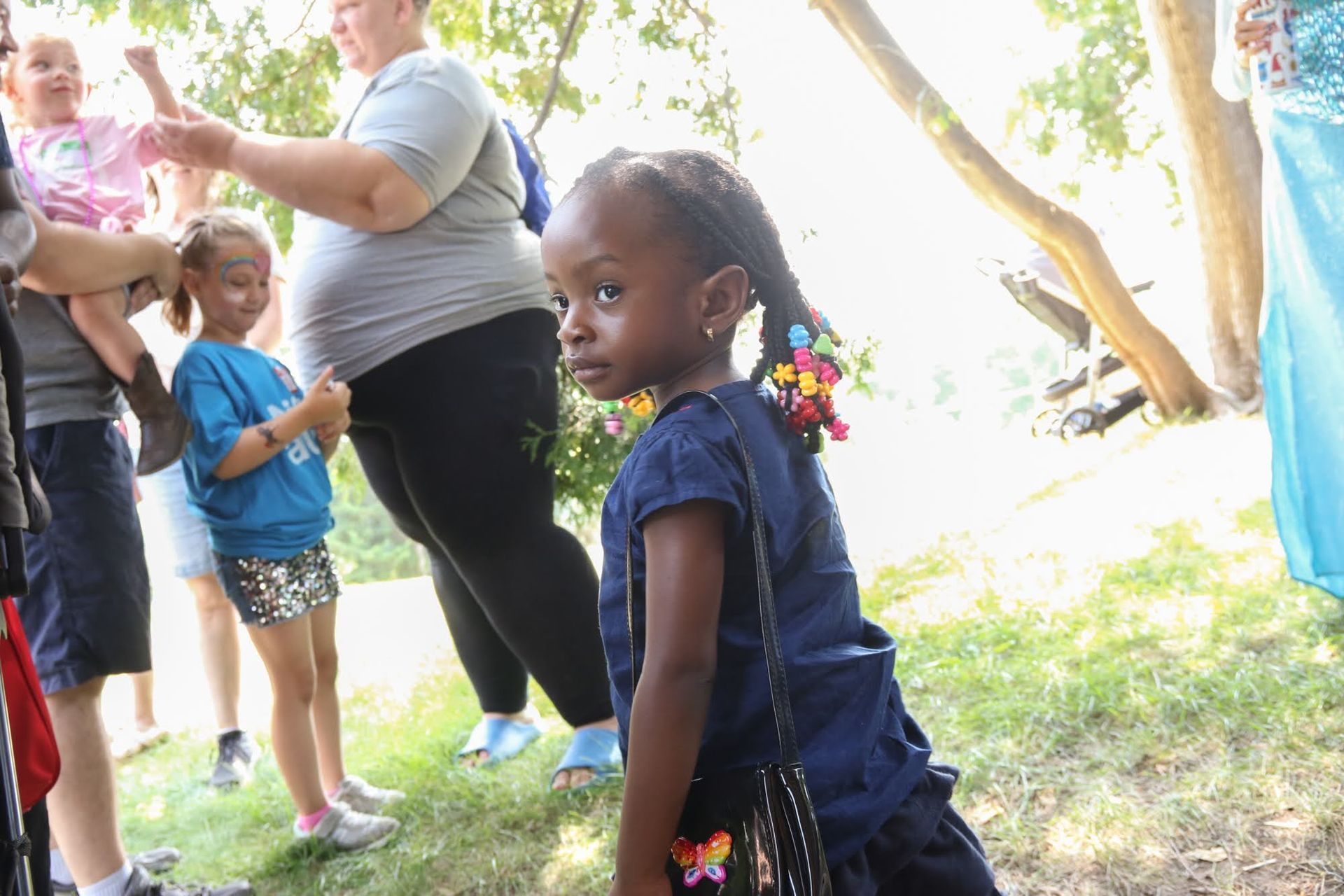 Girl with braids and colorful hair ties looks off-camera. People are nearby outdoors.
