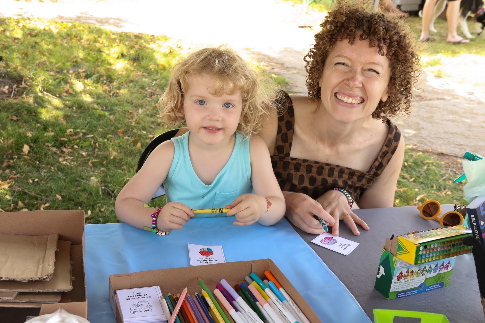 Woman and child smiling at a table with art supplies. Blue tablecloth. Outdoors.