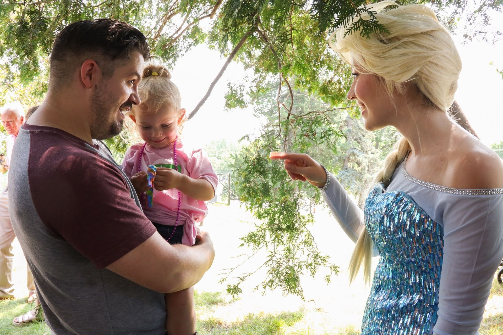 Man holding a child meets a costumed Elsa. Elsa points, smiling. Outdoors, sunny.