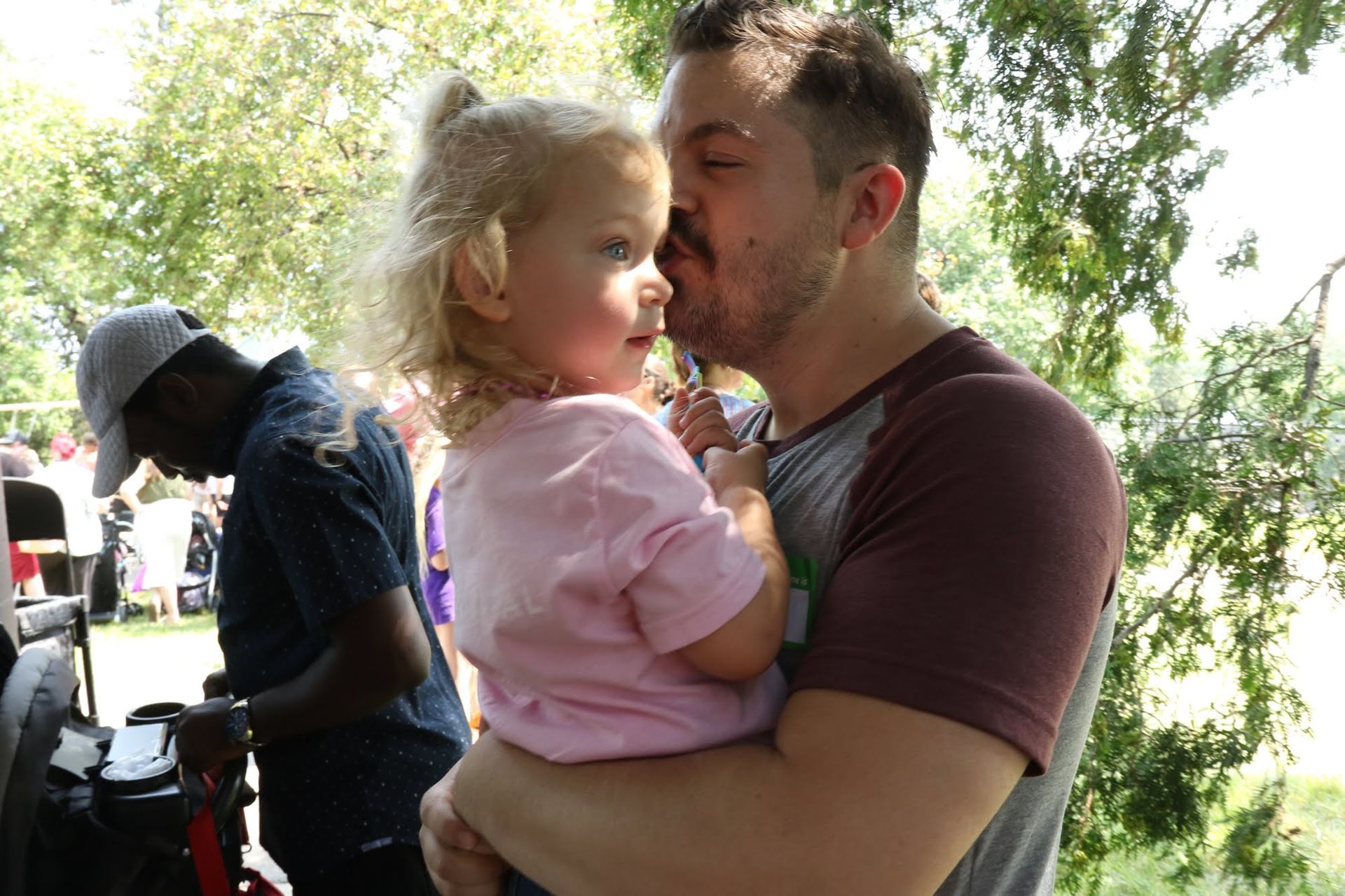 Man holding a toddler, kissing the child's cheek. Both are outdoors with other people in the background.