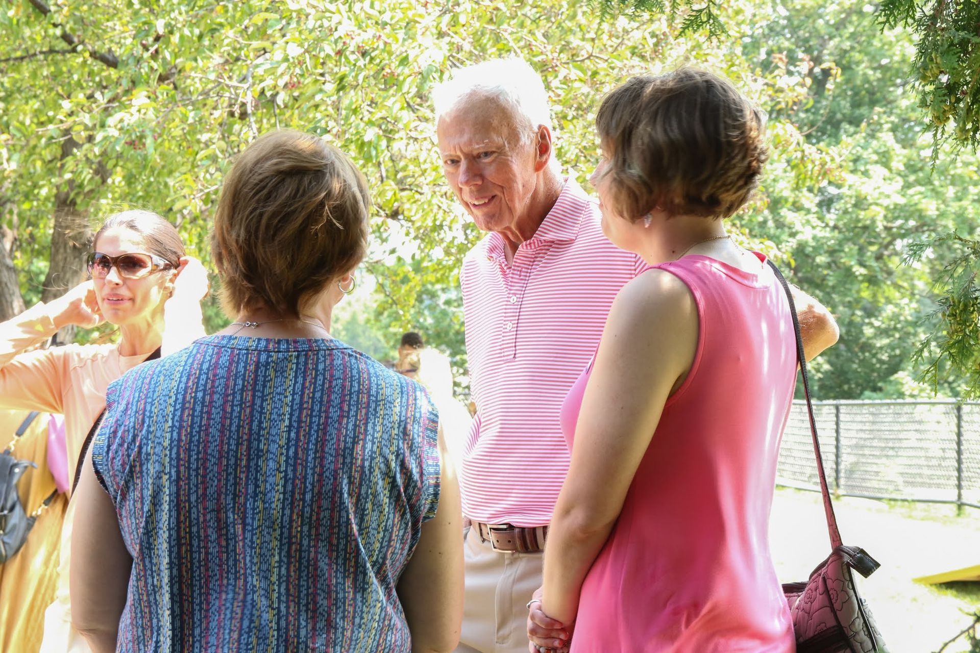 People conversing outside under trees. Man in pink shirt, women in various summer outfits. Sunlight.