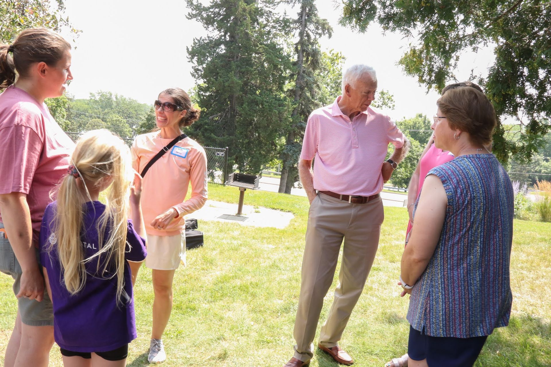 Group of people standing outside on a grassy area, talking. A man in pink shirt, other women. Girl in purple shirt.