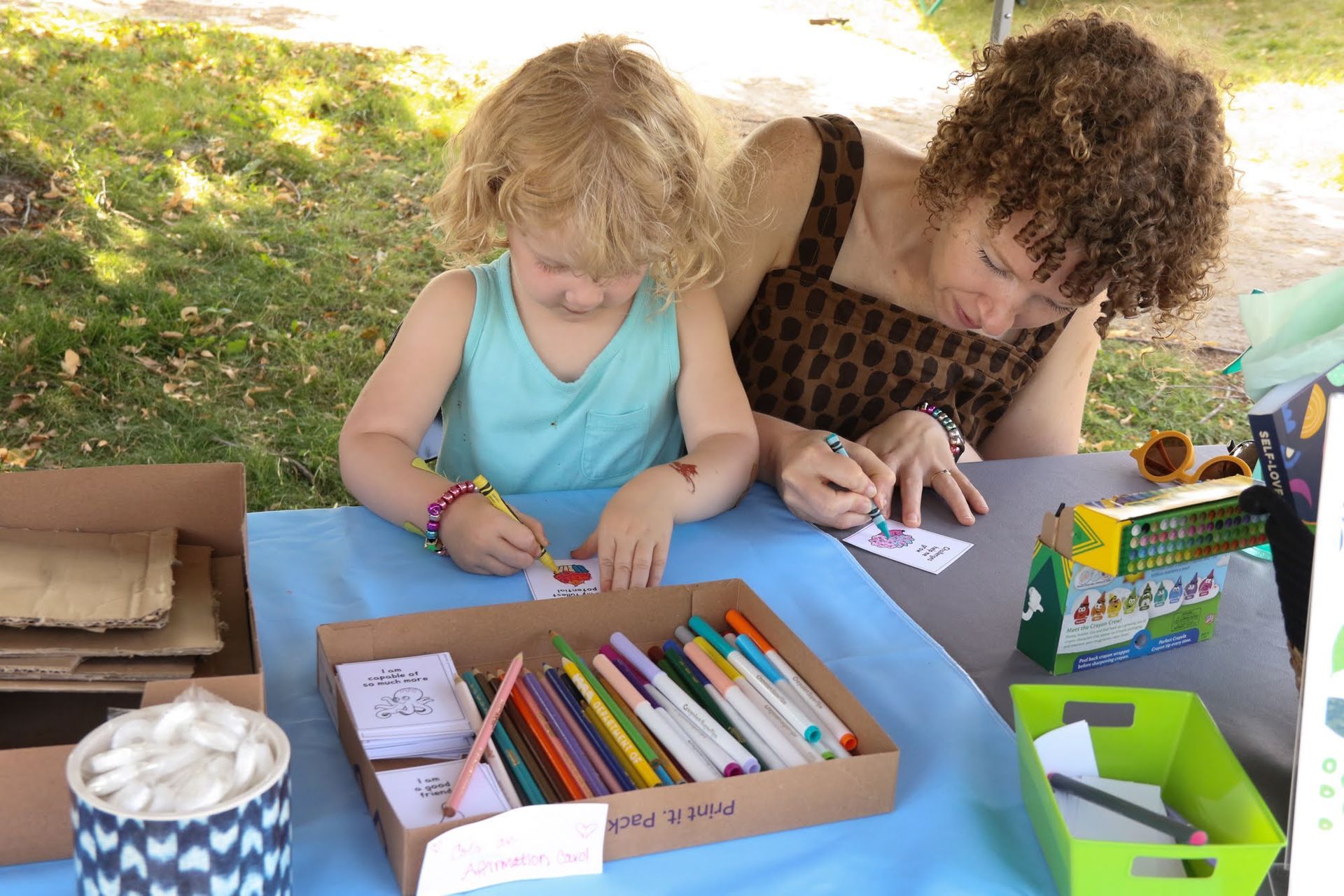 A person and child are coloring at a table with markers and cards outdoors on a sunny day.