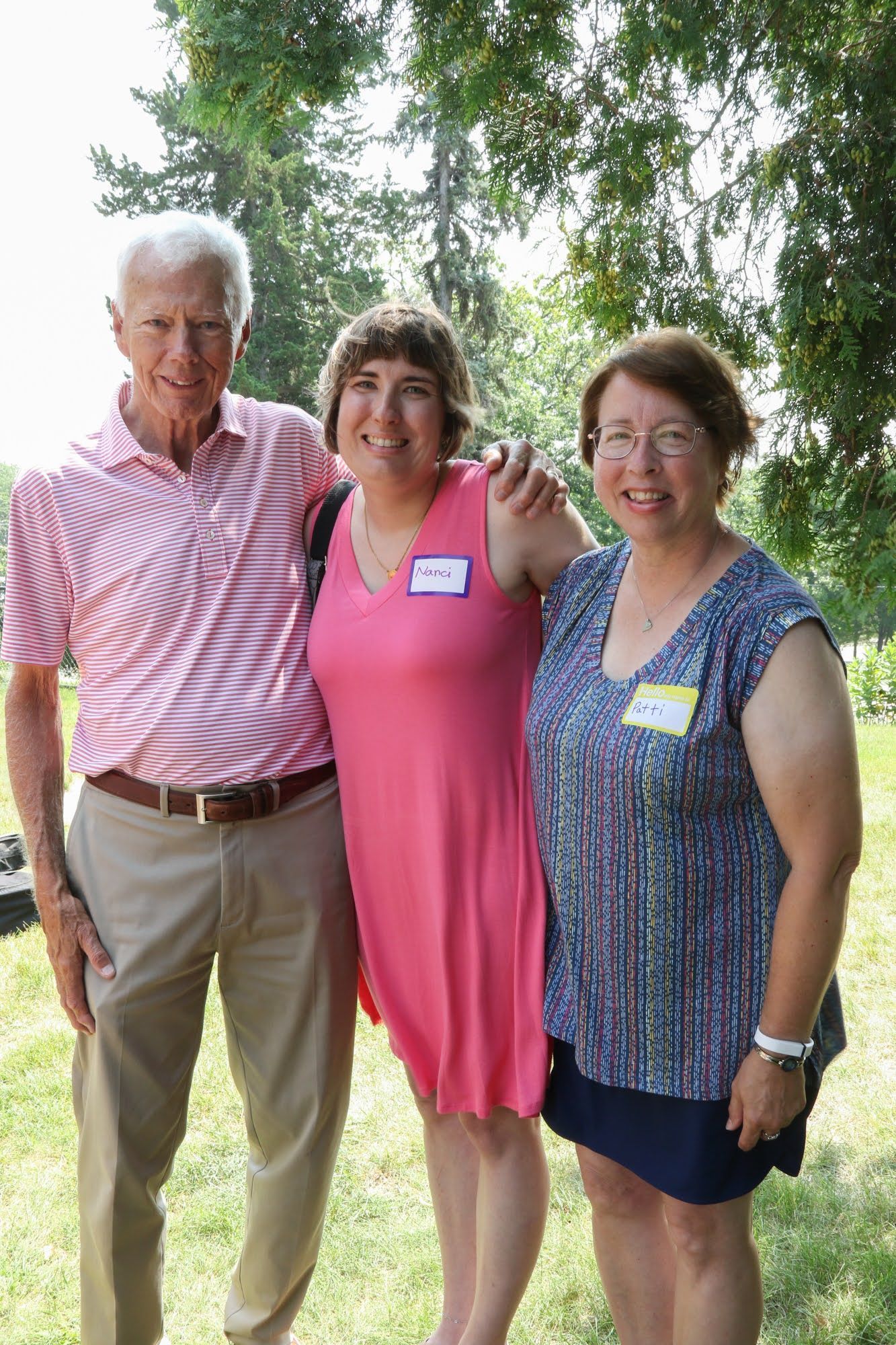 Three people, posing outdoors. A man in a pink shirt and khaki pants stands with two women, one in pink, one in blue.