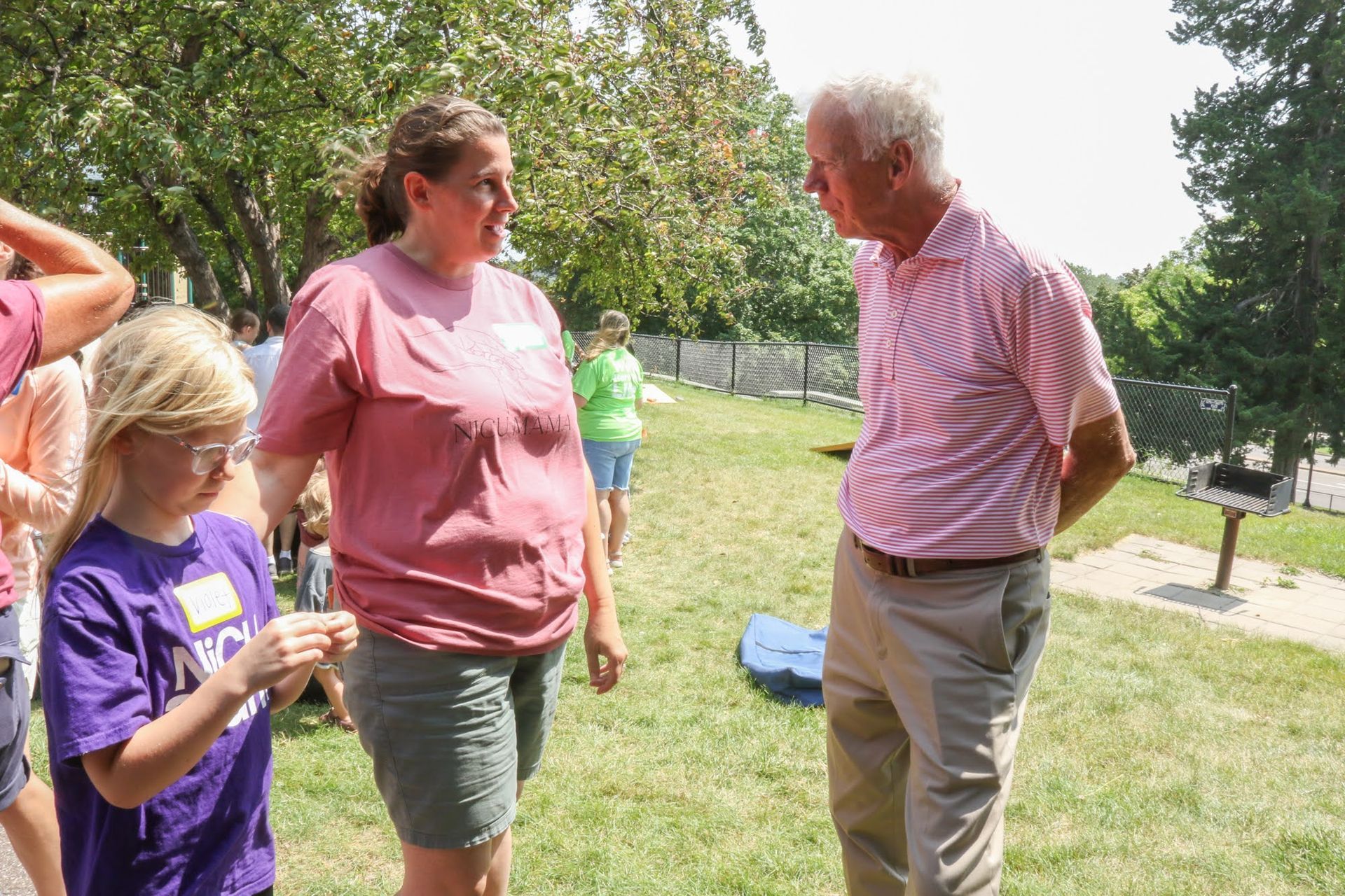 A man in pink shirt speaks with a woman in pink shirt, a girl in a purple shirt looks on in a park.