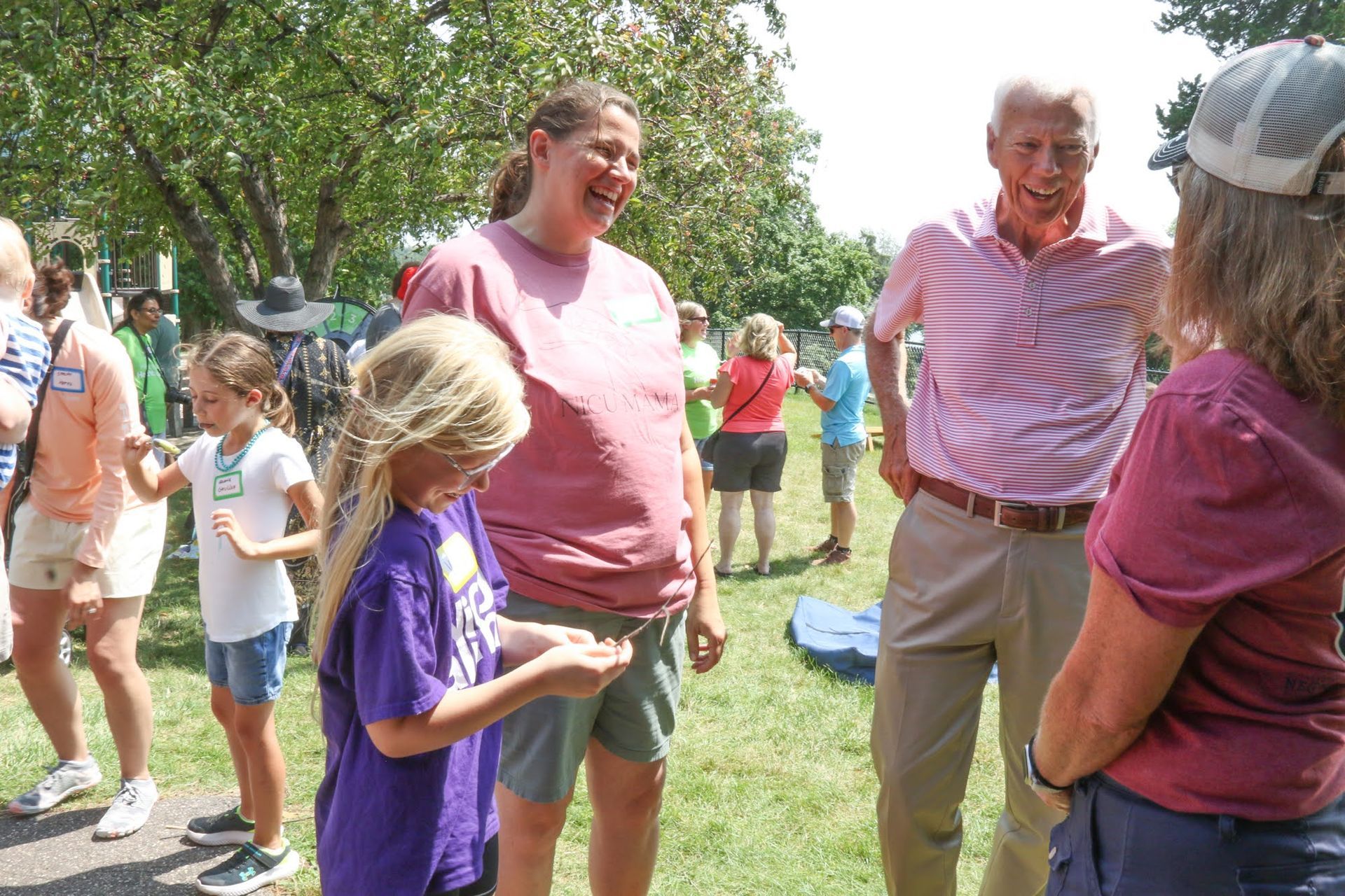 People outdoors talking. Woman in pink shirt smiles at girl holding something, man in pink and white striped shirt also smiles.