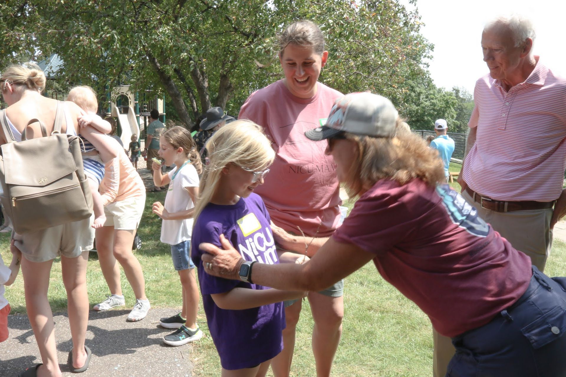 Woman adjusts a young girl's name tag outdoors, surrounded by people; sunny day.