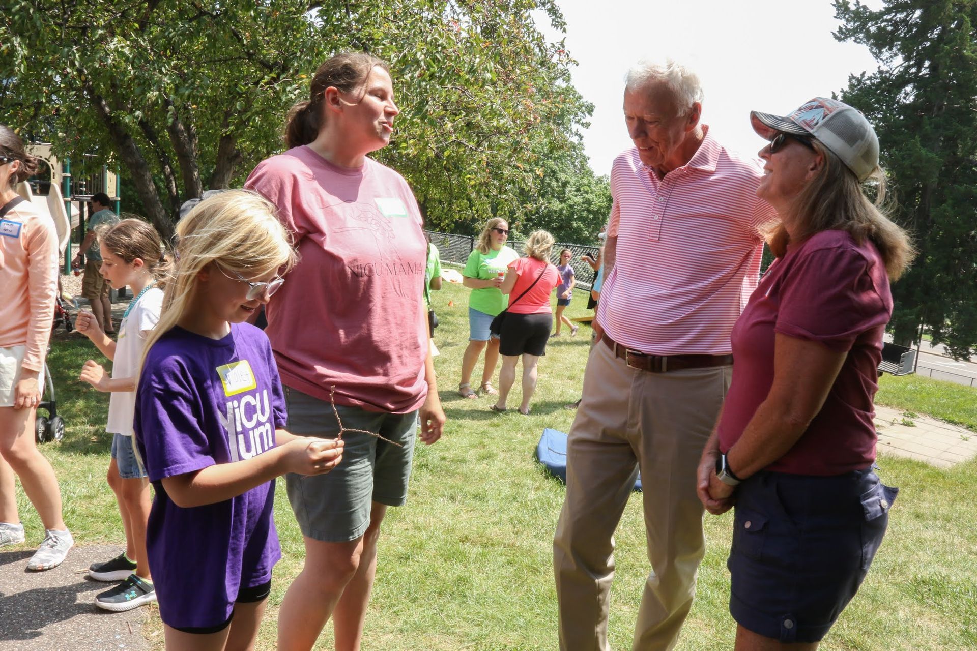 A group of people outdoors, talking. A young girl shows a string. A man in pink stripes, and two women in maroon shirts are present.