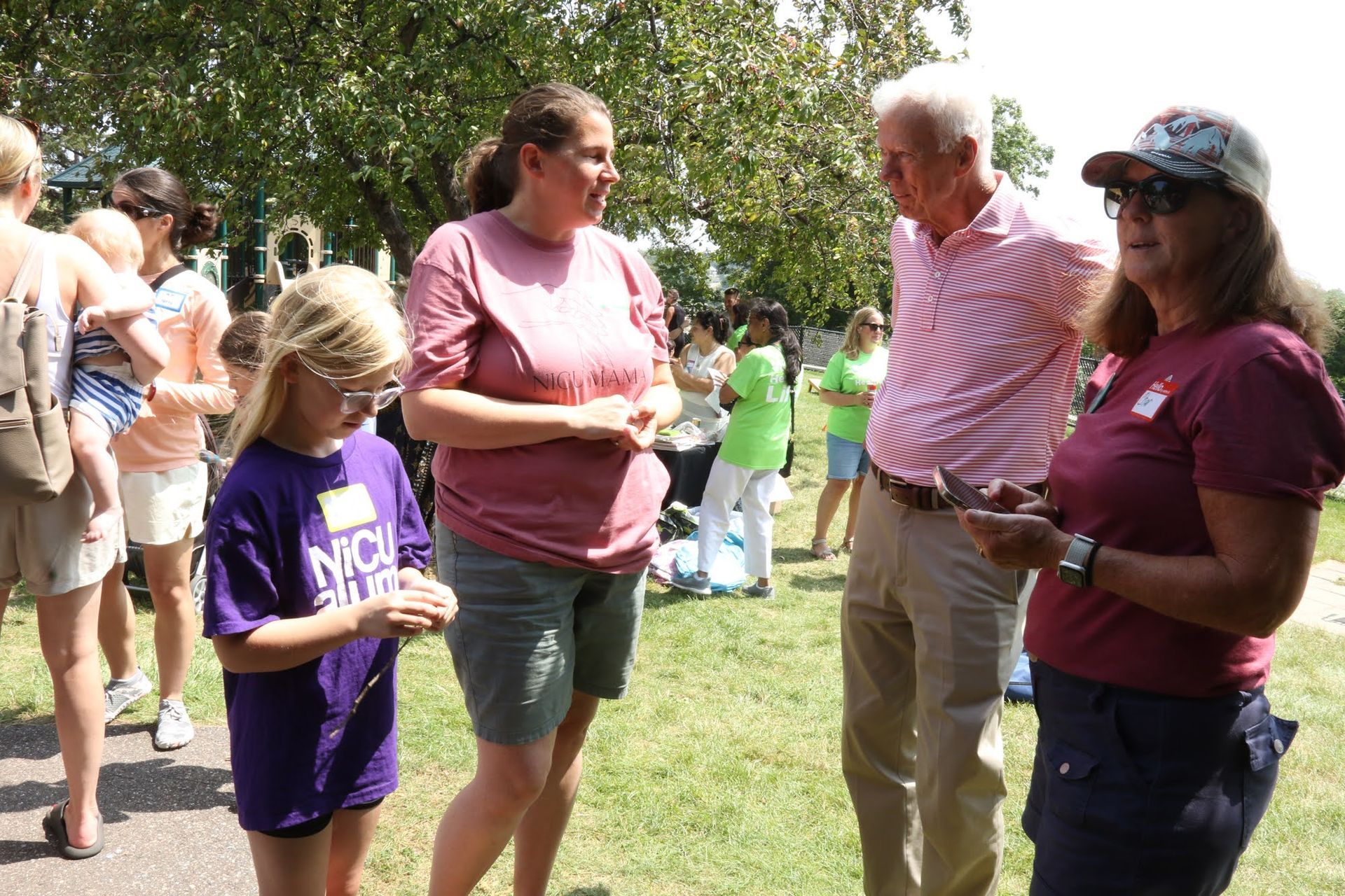 People talking outside on a sunny day. Girl in purple shirt bows head. Woman in cap holds a phone.