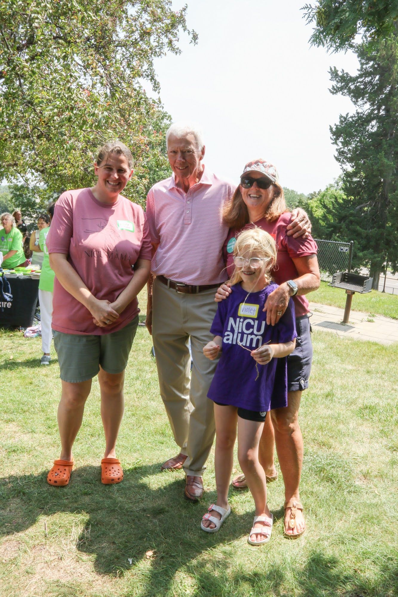 Four people pose outdoors. A man in pink shirt stands with three women. One child wears a purple shirt.