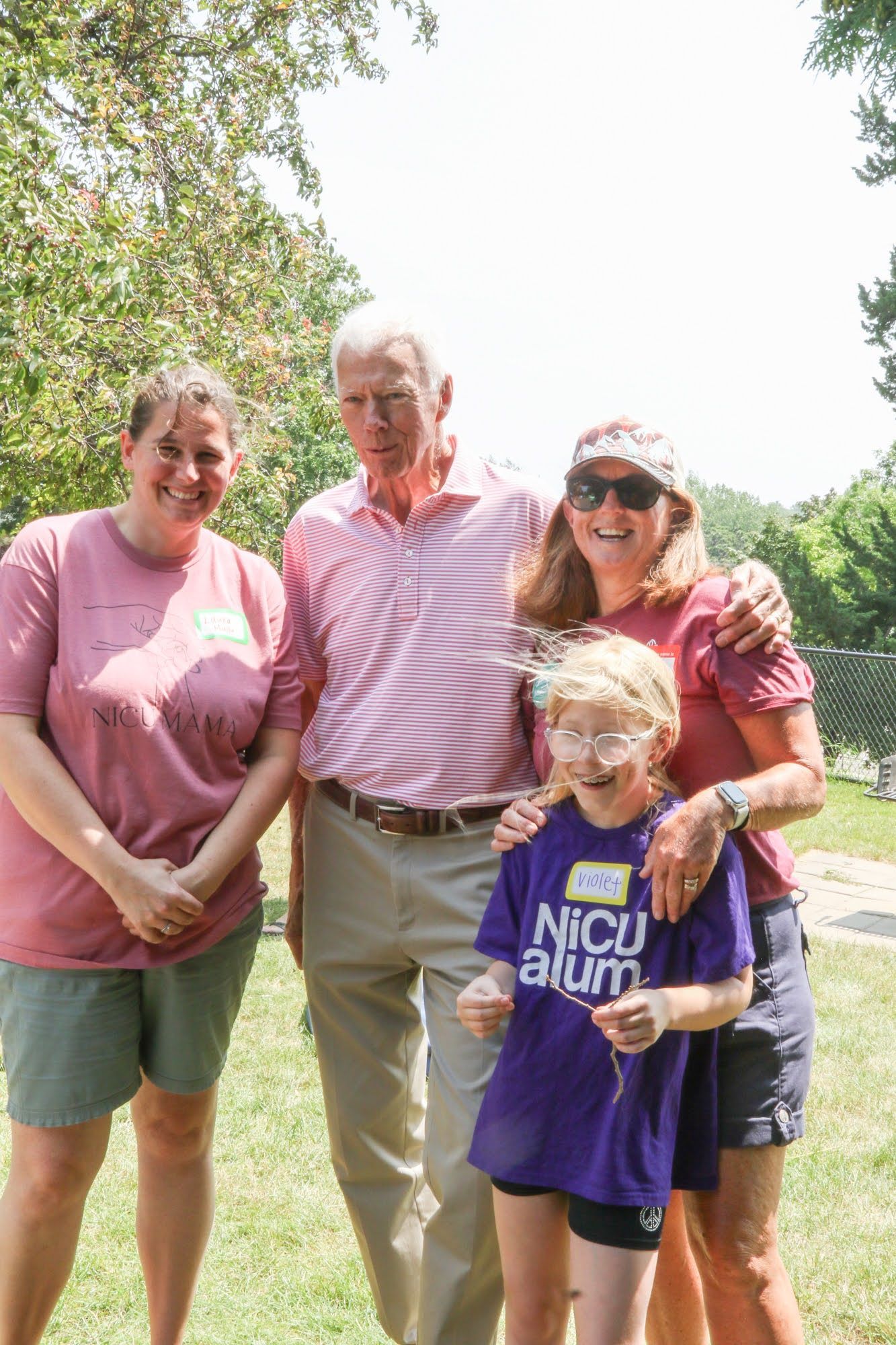 Four people pose outdoors: a man, a woman, a girl, and another woman. All wear casual clothes, smiling.