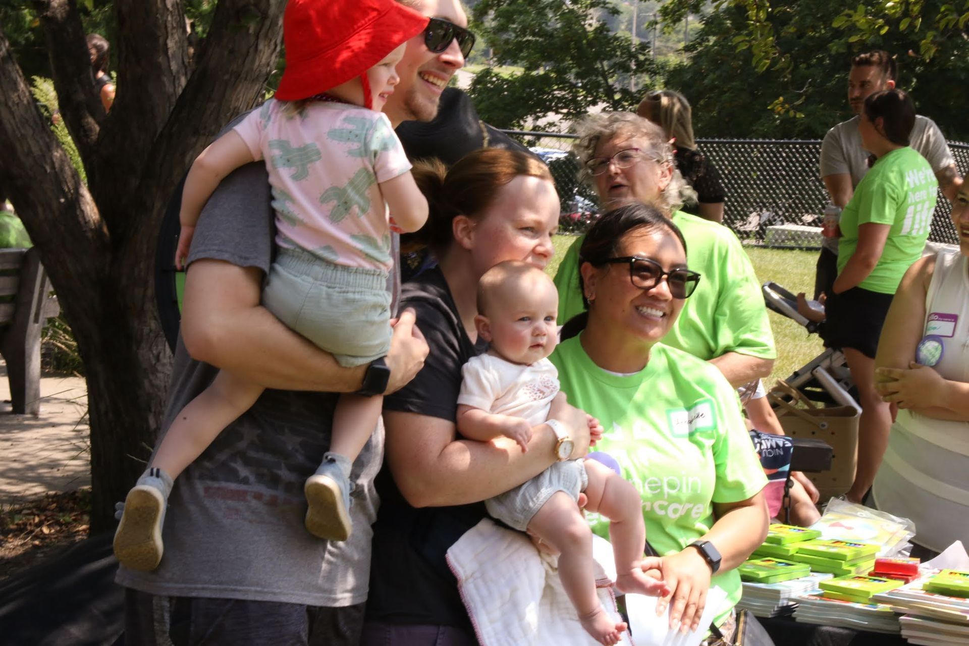 Group of people with babies, smiling outdoors, some wearing green shirts.