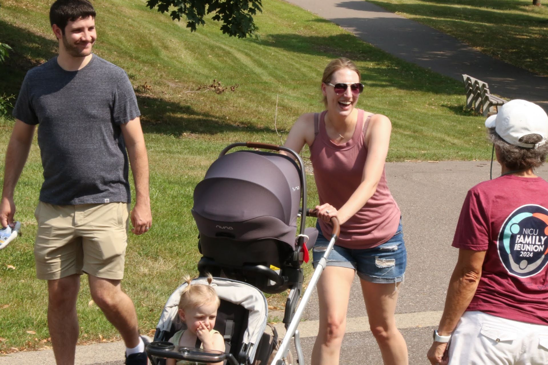 People walking on a path in a park; a woman pushes a stroller, smiling.