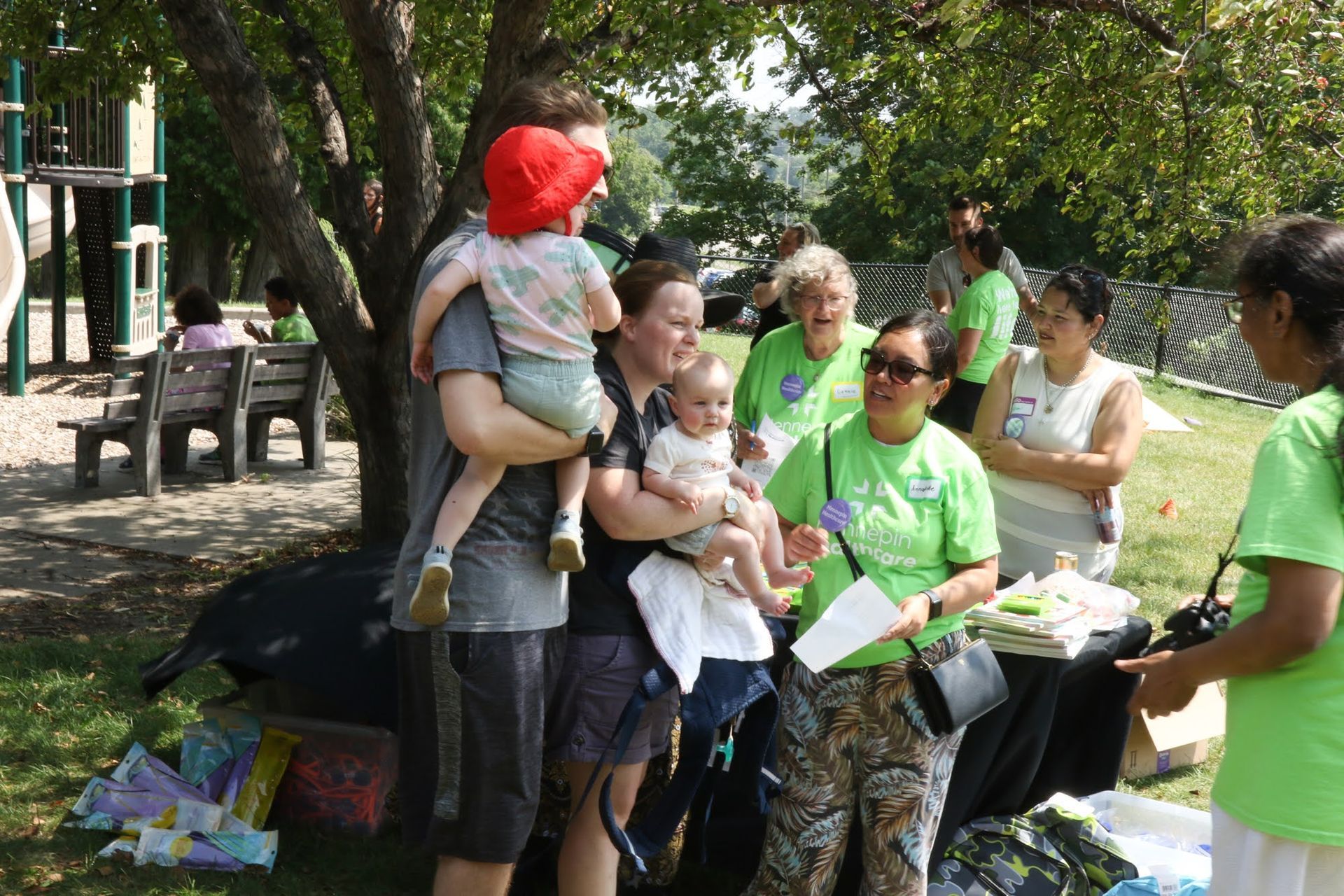 People at an outdoor event with babies, some wearing green shirts. Playground in background.