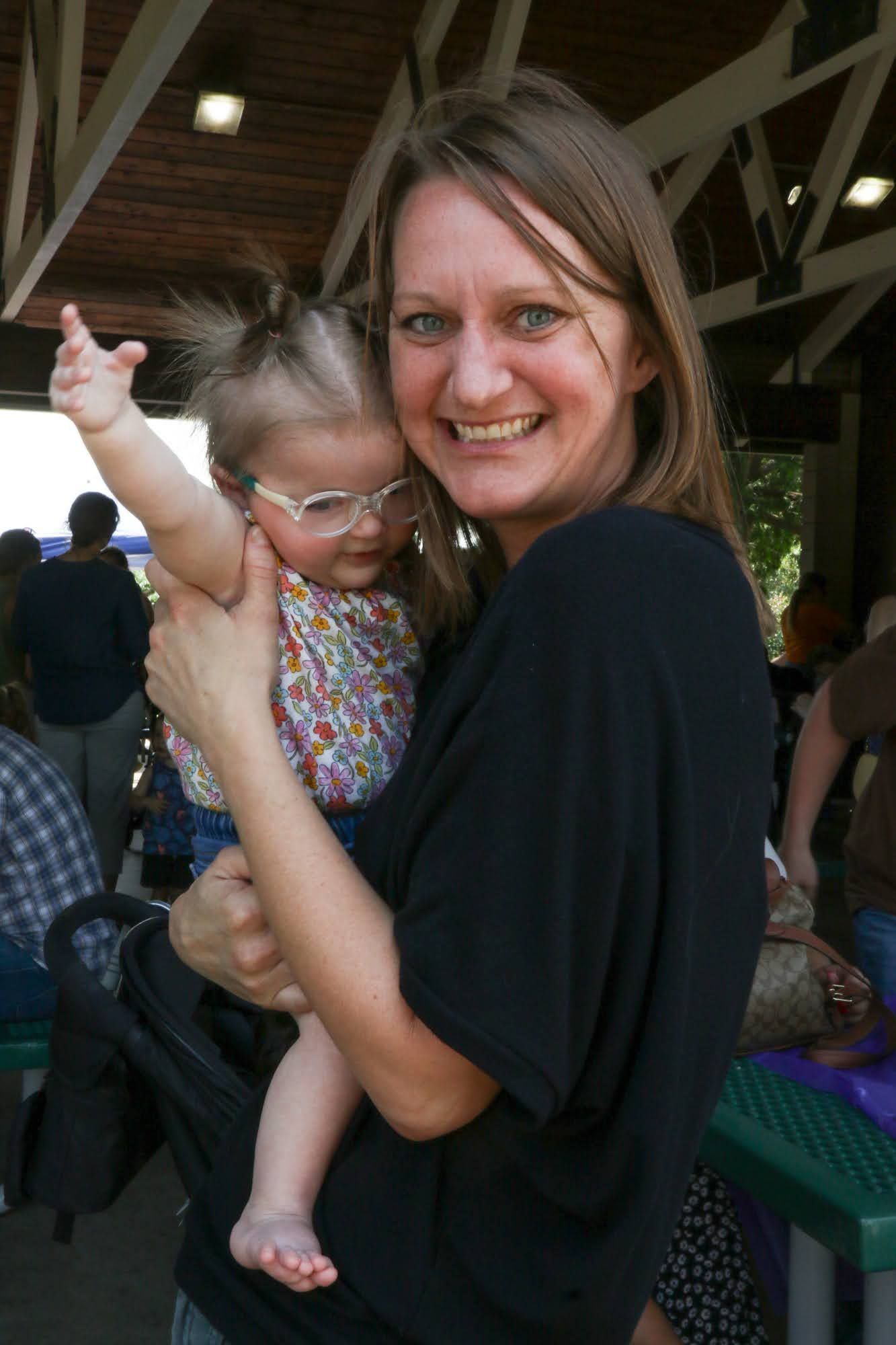 Woman holding a baby wearing glasses, outdoors. The woman smiles, baby's arm raised.