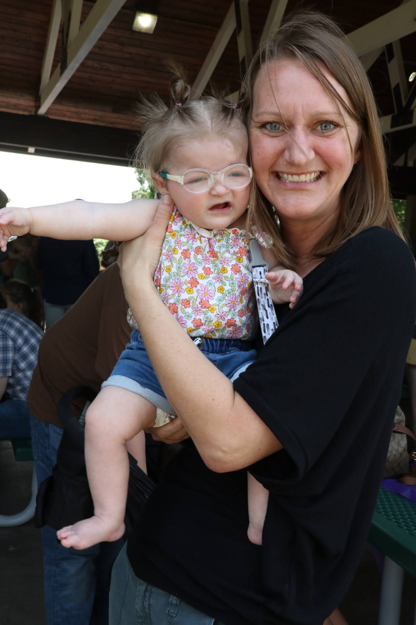 Woman holding a baby wearing glasses in an outdoor setting; both smiling.