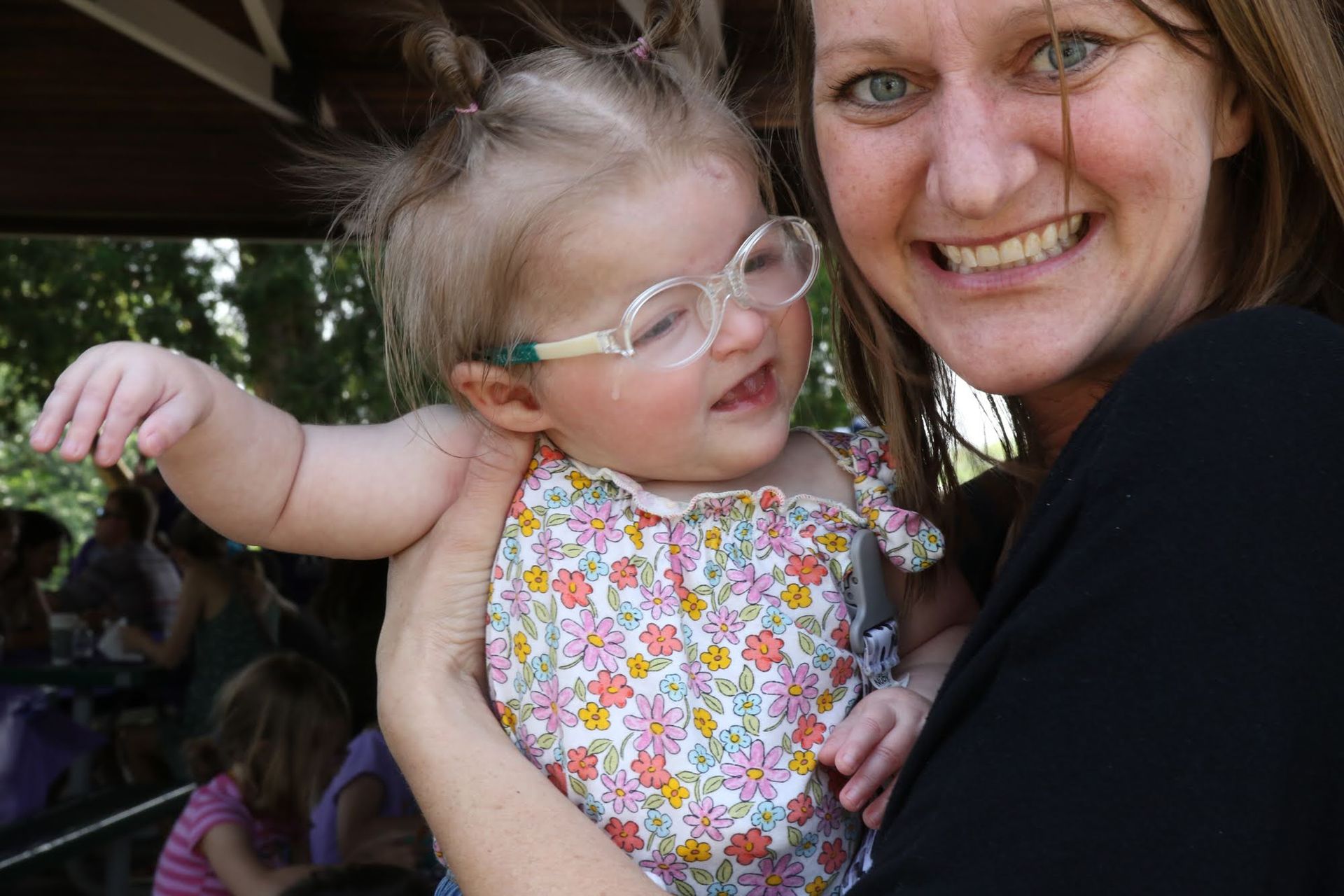 Woman holding a smiling baby wearing glasses and a floral dress. They are outdoors.
