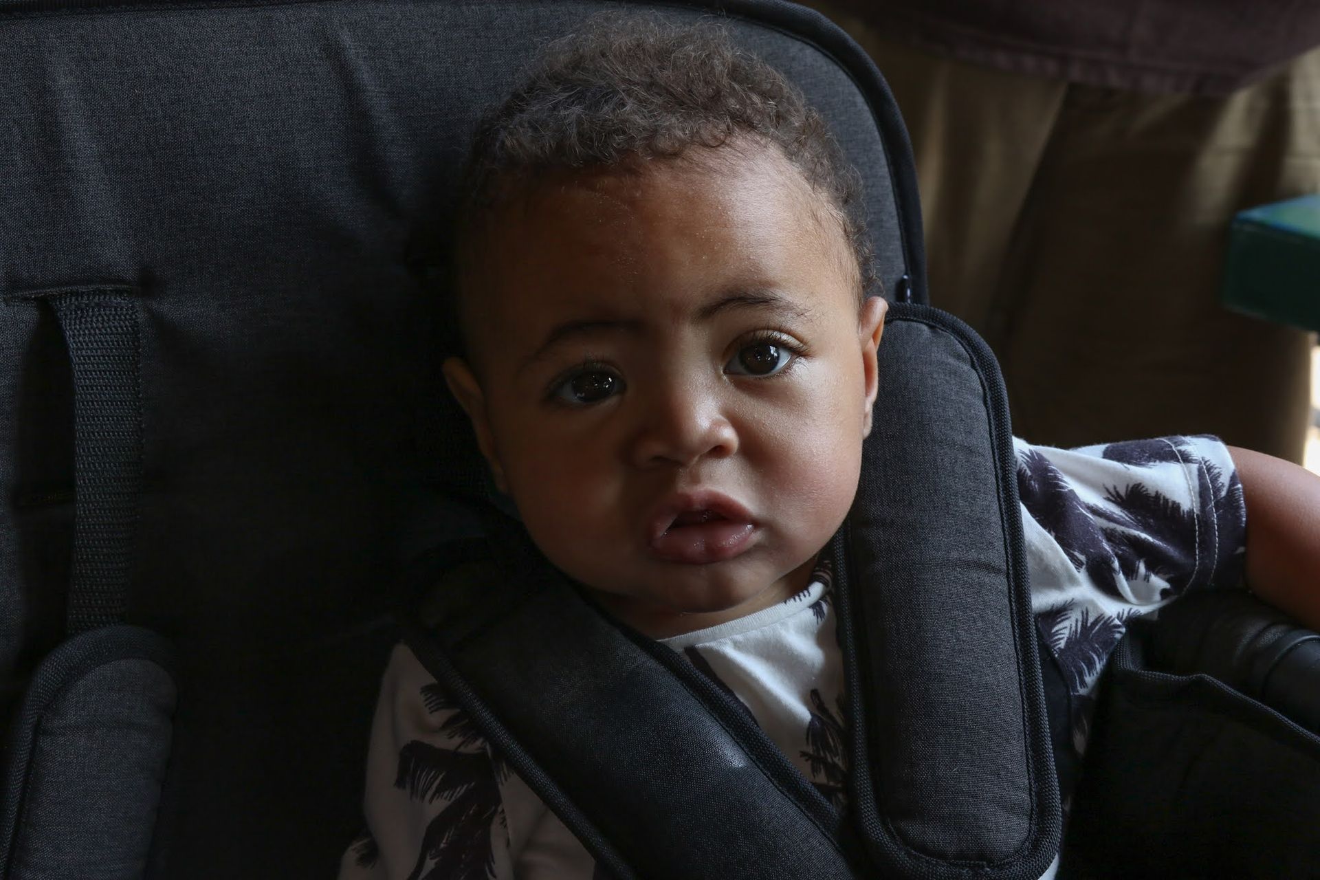 Baby in a car seat, looking directly at the camera with a neutral expression. Black seat belts and grey-white shirt.