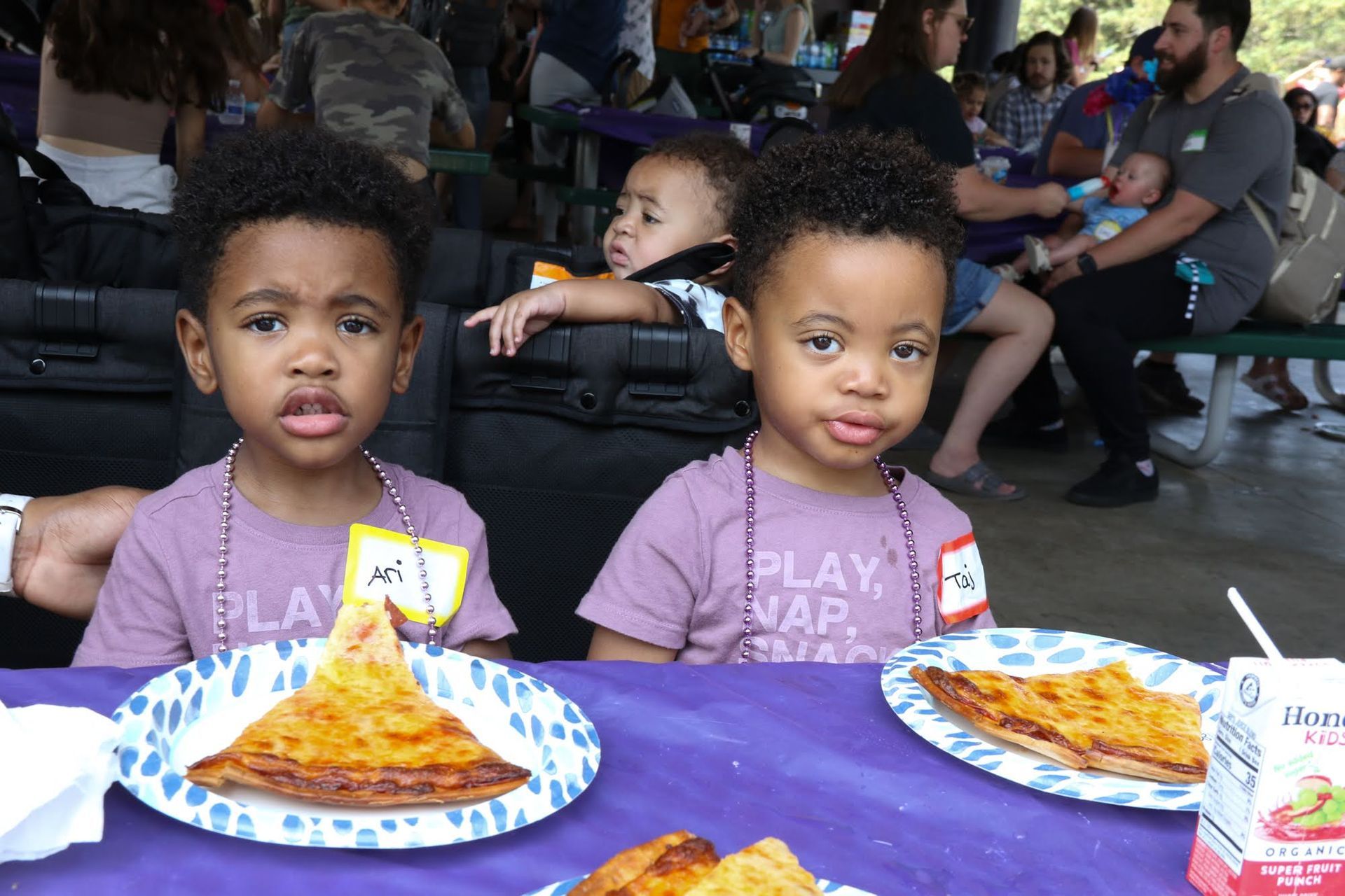 Two young children eating pizza outdoors. They are sitting at a table with other people in the background.