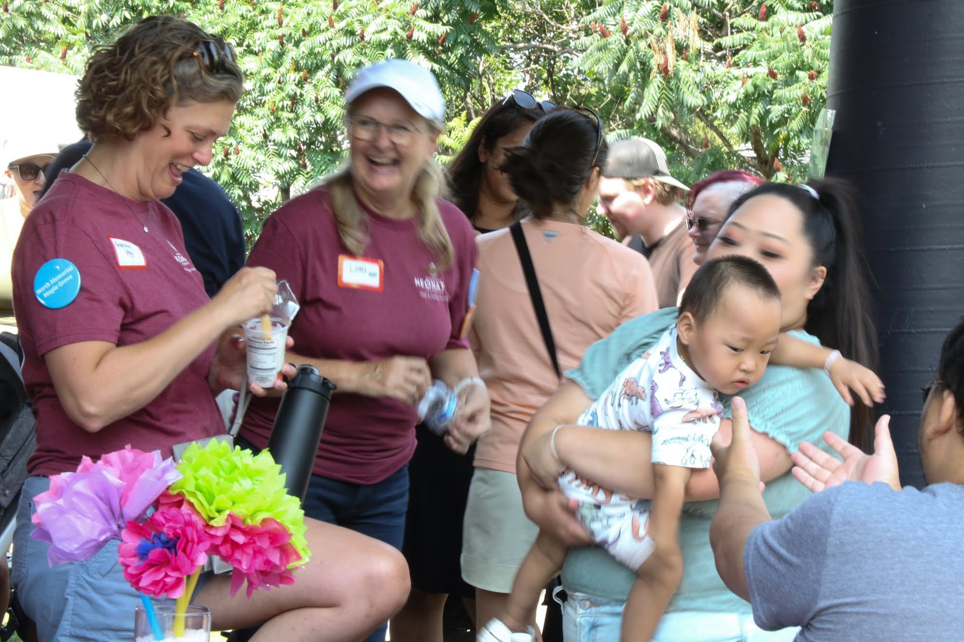 People at an outdoor gathering. Woman holds a child; two women laugh. Flowers in front, other people in background.