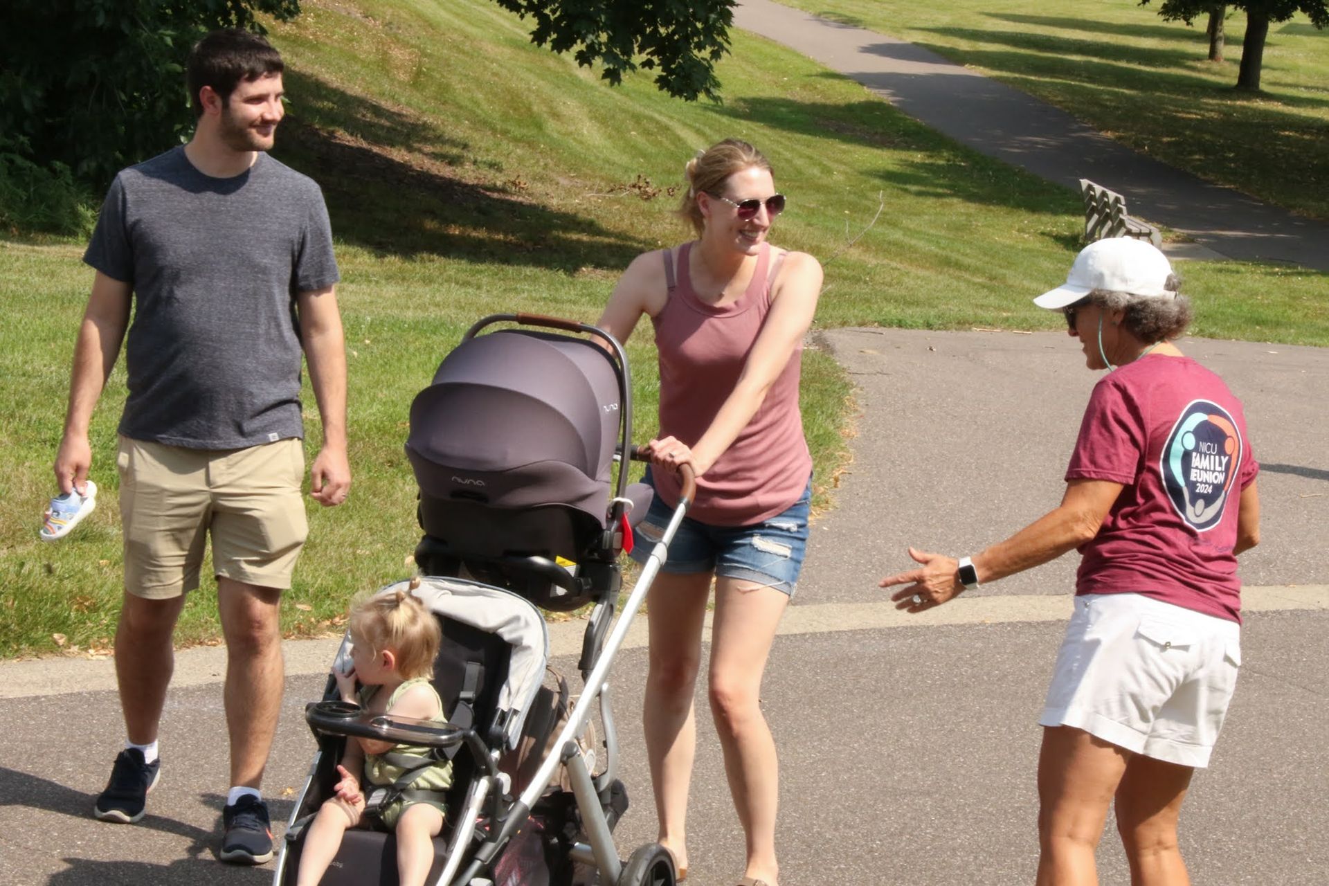 Man and woman with stroller and child walking on a path, greeted by another woman wearing a visor. Sunny outdoor setting.