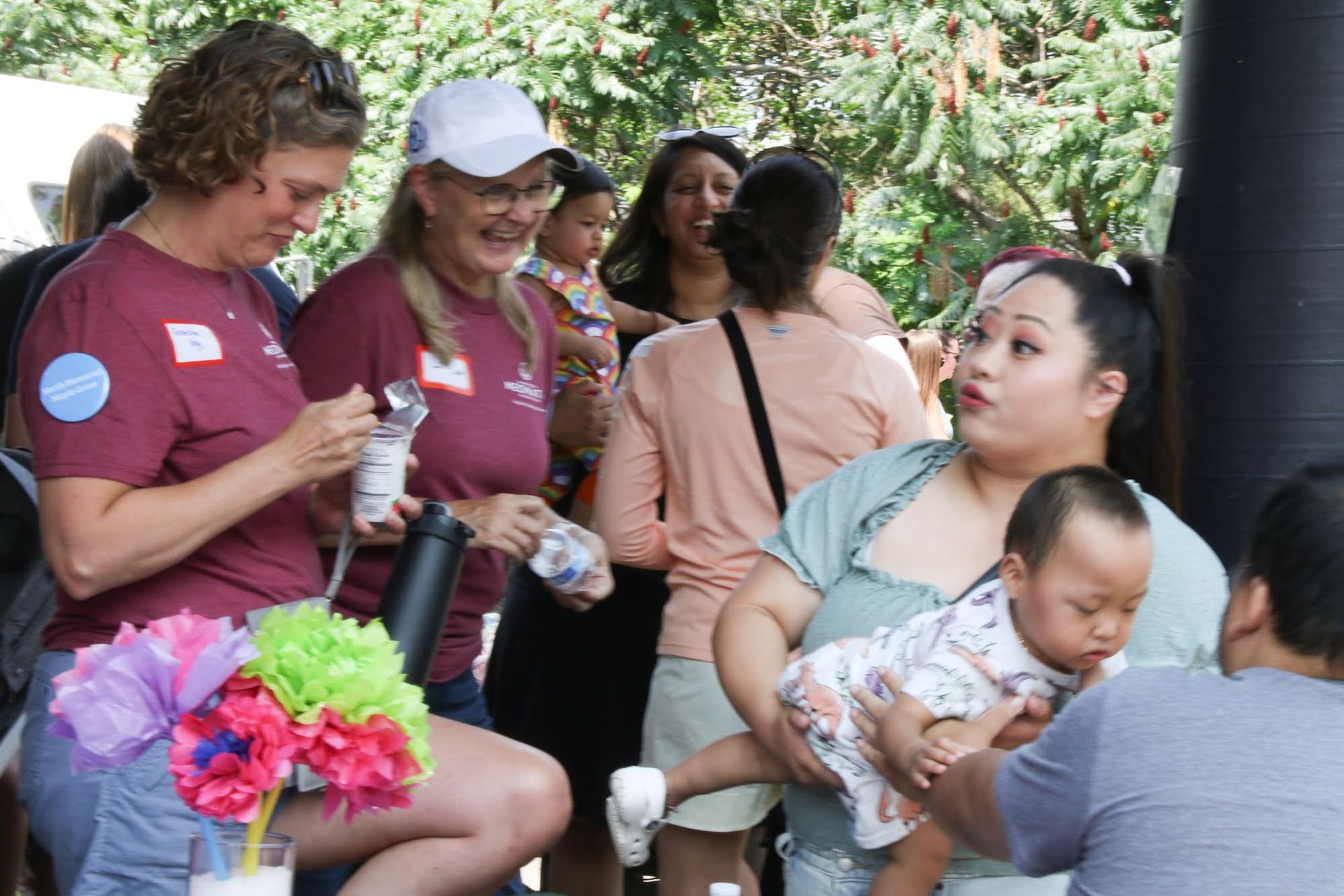 People at an outdoor event, some holding babies, with colorful decorations.