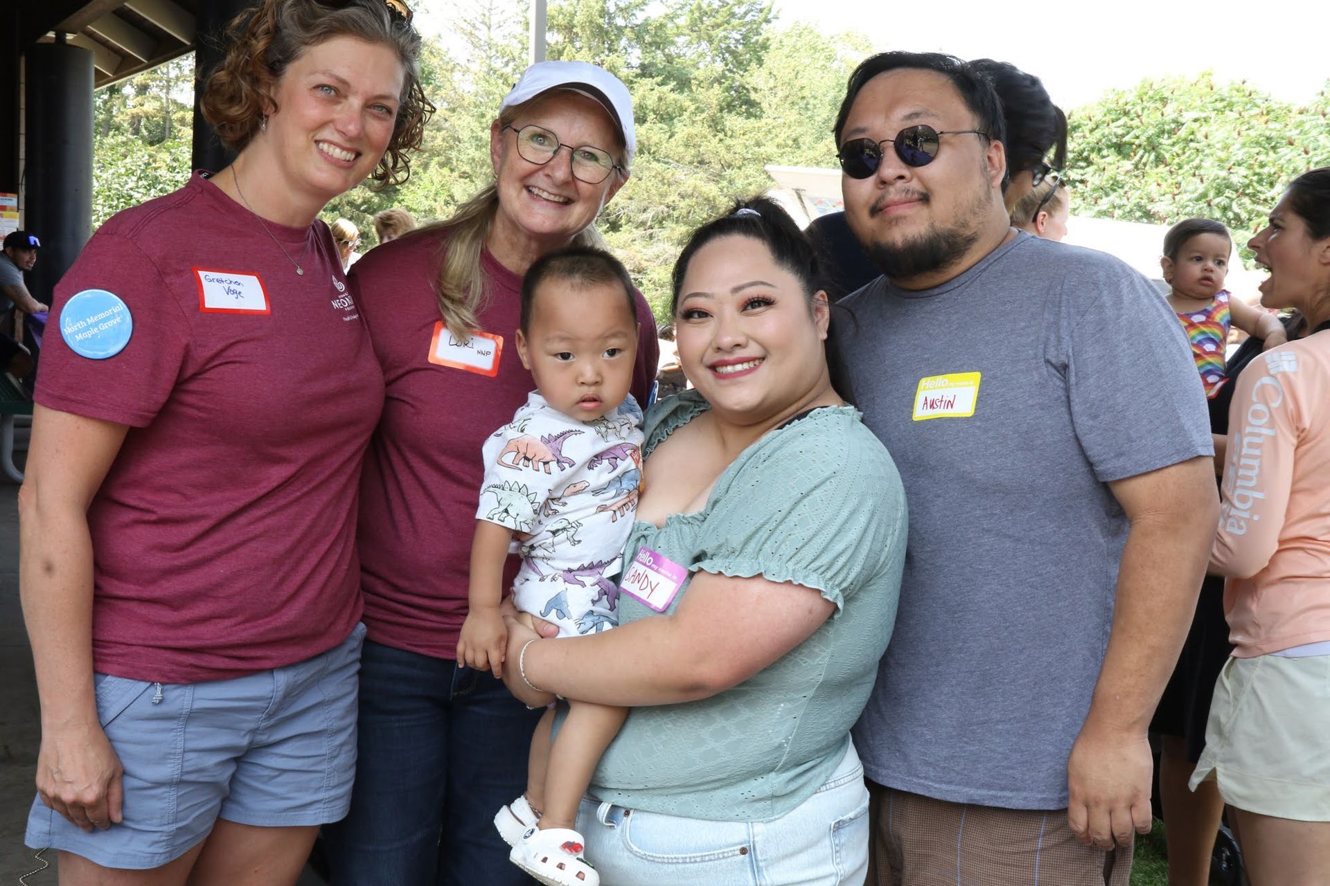 Group of five people smiling, one holding a baby. Outdoors, sunny.