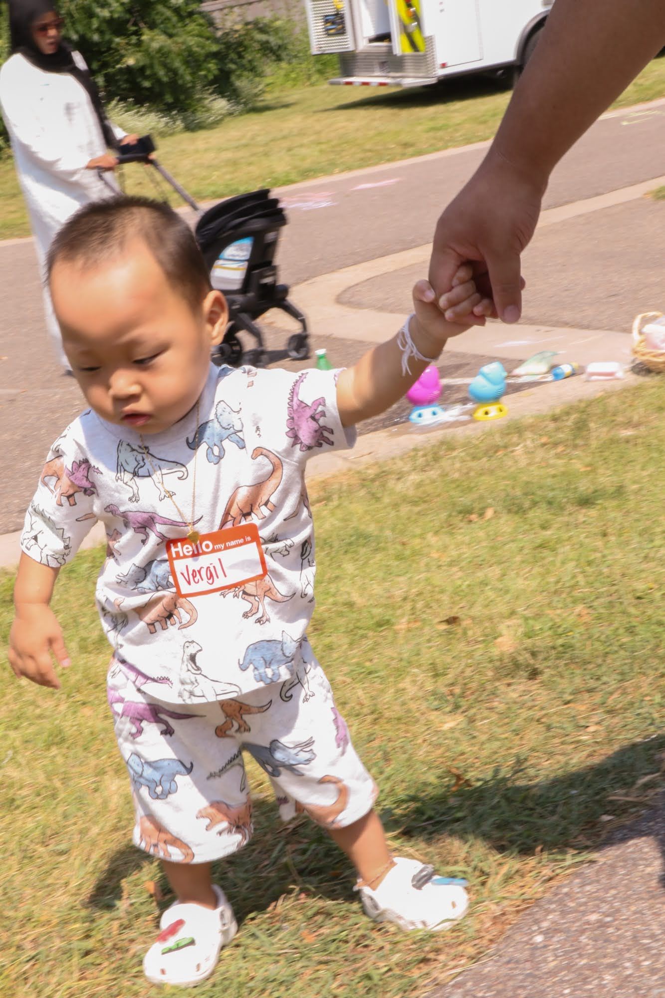 Child walking, holding an adult's hand, wearing dinosaur-print outfit and name tag, outdoors on grass.
