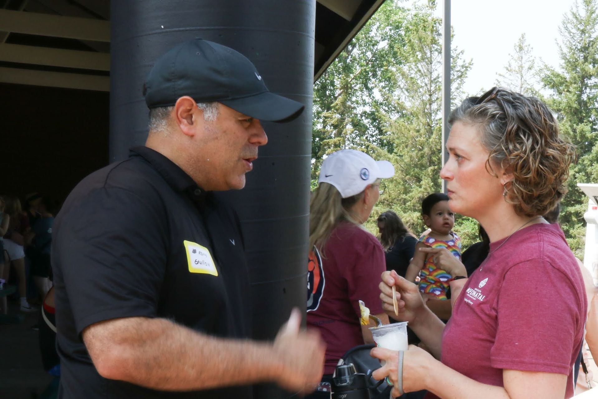 Man in black shirt and cap speaks to woman in maroon shirt outdoors.