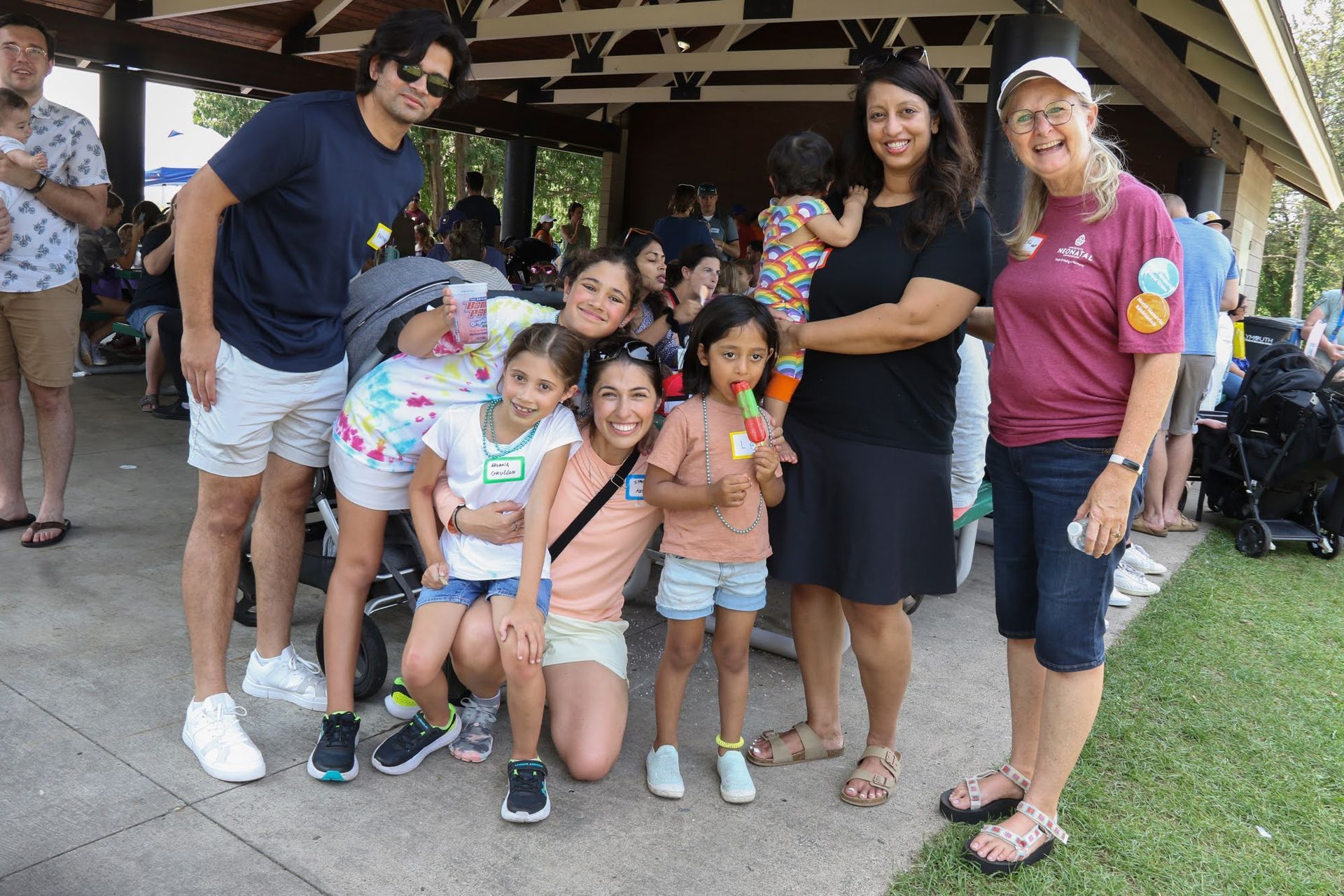 Group of people smiling for photo at outdoor gathering under a pavilion; some children holding treats.