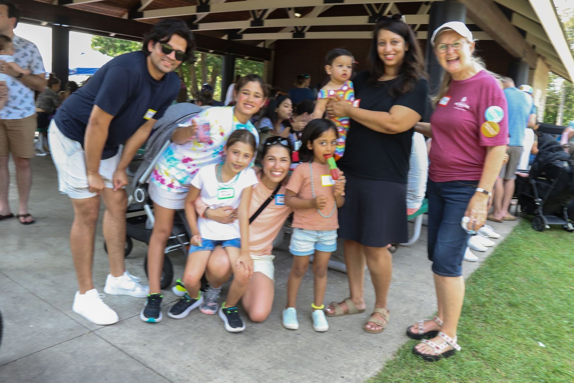 Group of people smiling, posing outdoors near a pavilion. Several children with adults, some holding treats.