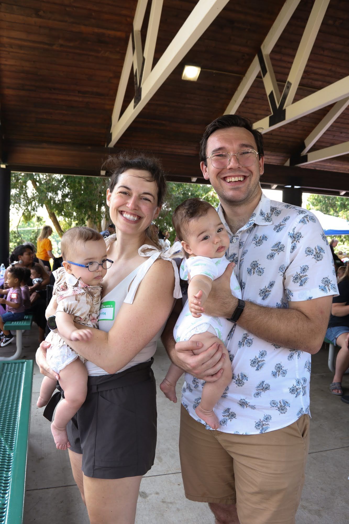 A smiling couple holds twin babies. They stand outdoors under a shelter, the babies wearing matching outfits.