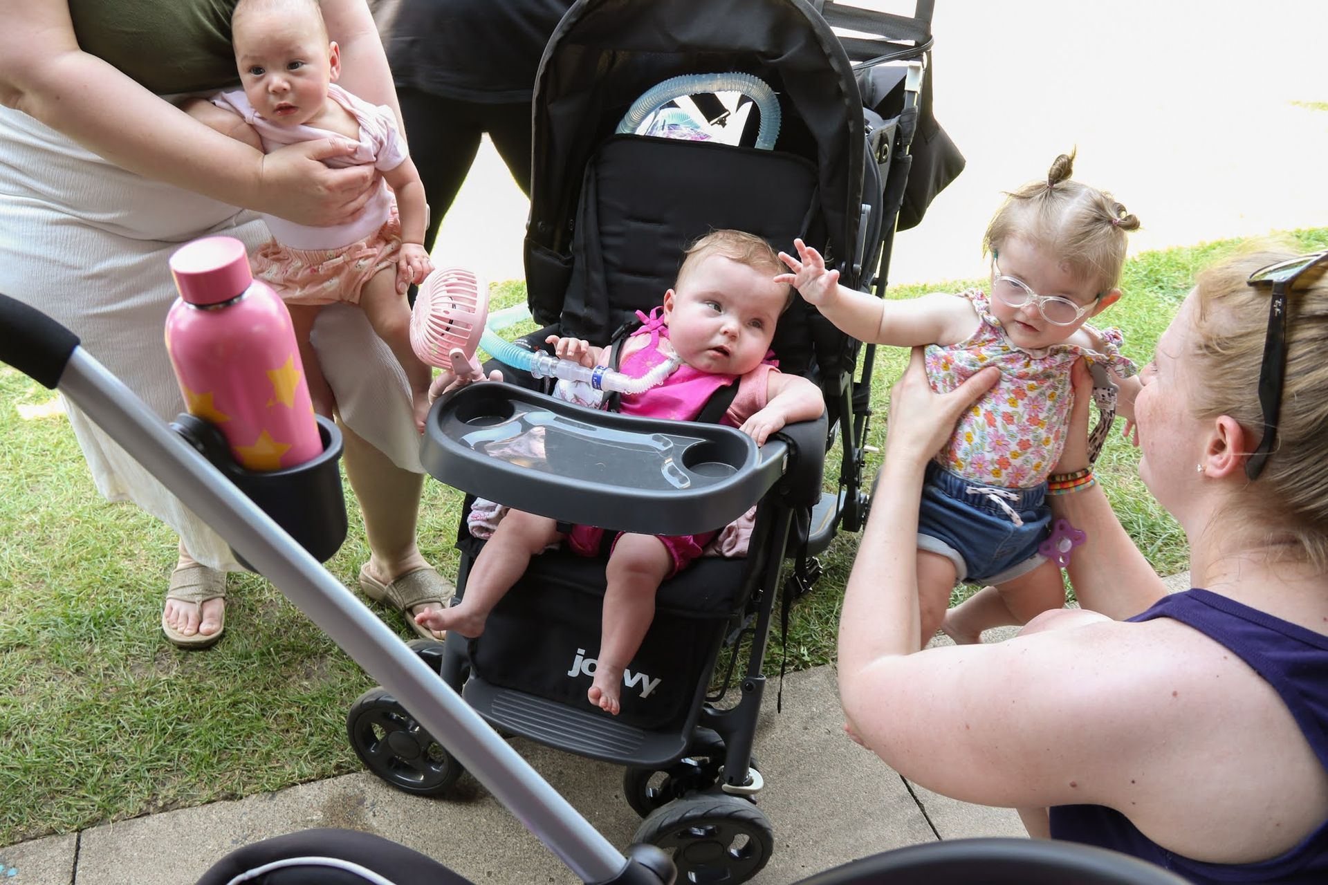 Four babies, two in a stroller, two held by adults, outdoors. Pink water bottle in stroller cup holder.