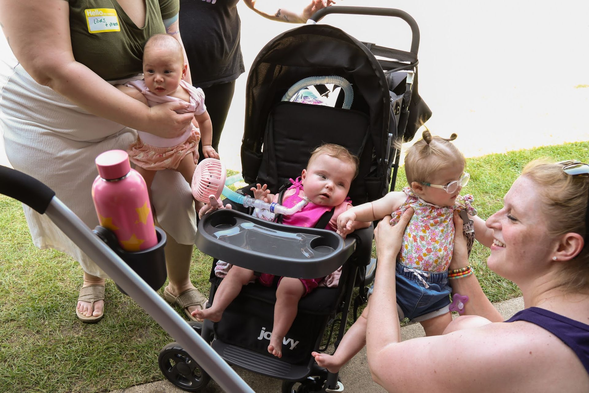 Babies with caregivers near a stroller. One baby in stroller, others held. Outdoors.