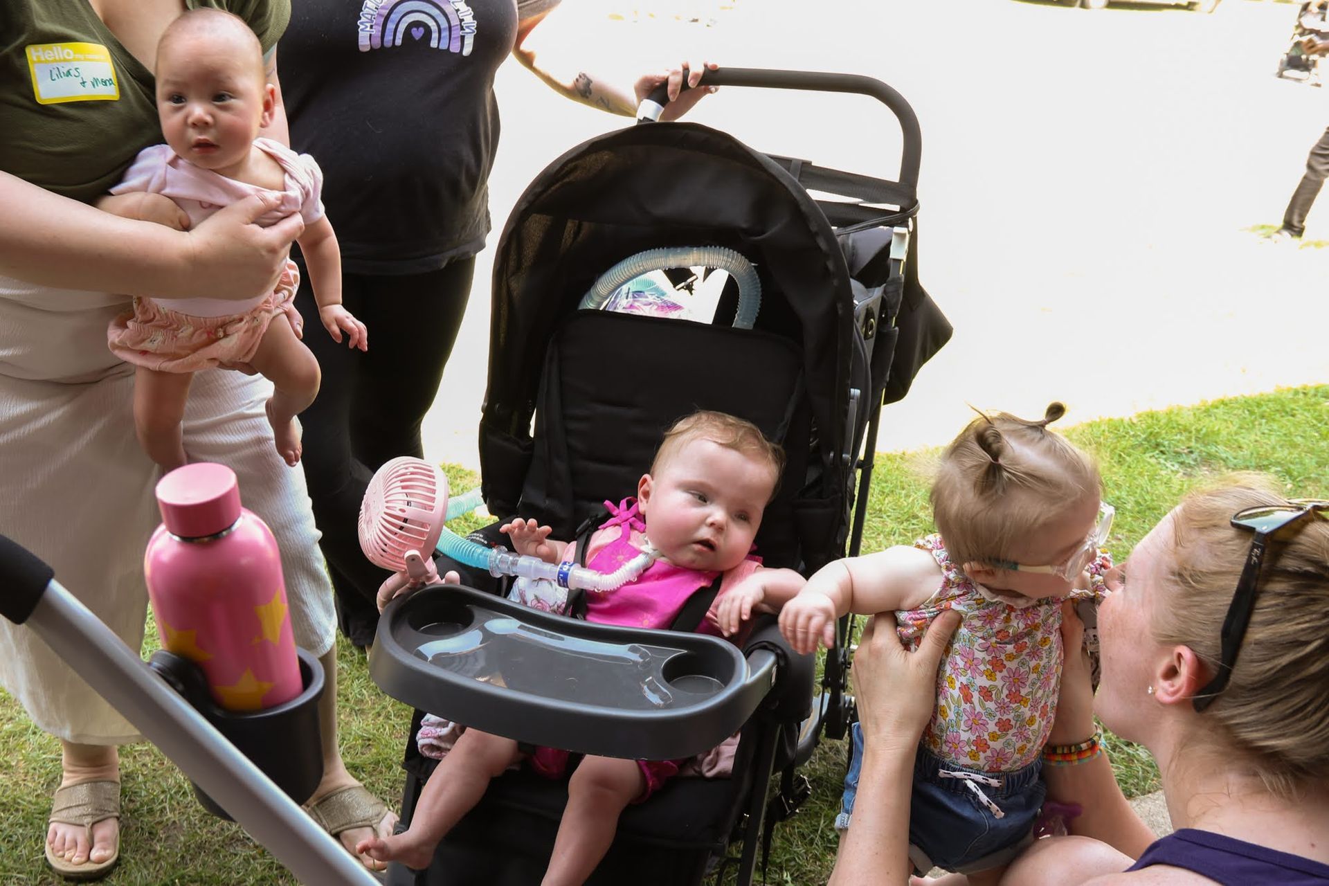 Four babies with their caregivers, two babies in stroller, the other two held. Outdoors.