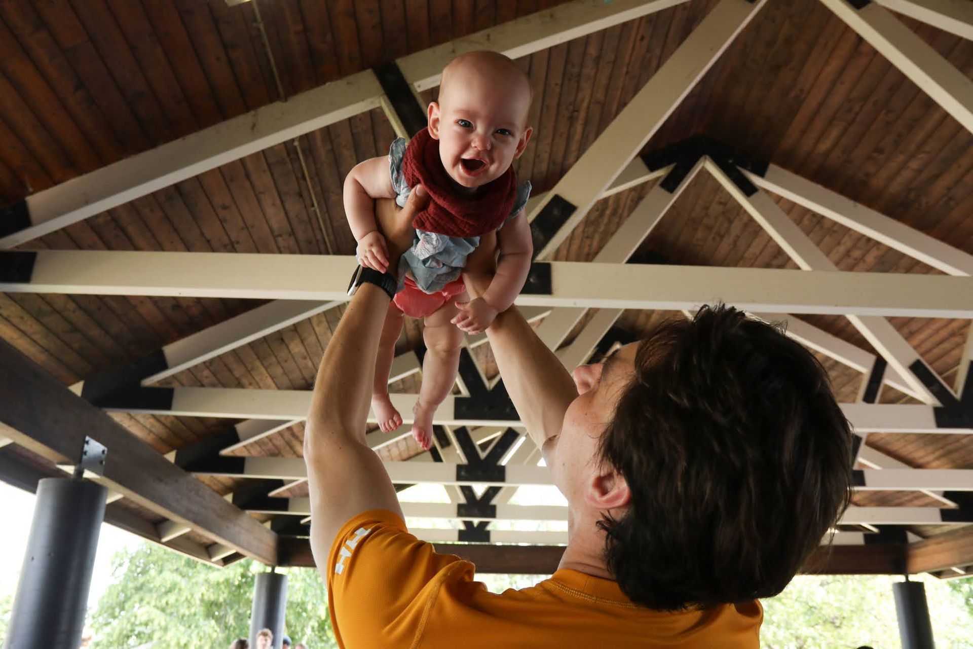 A person in an orange shirt holds a baby up in the air under a wooden gazebo roof. The baby is crying.