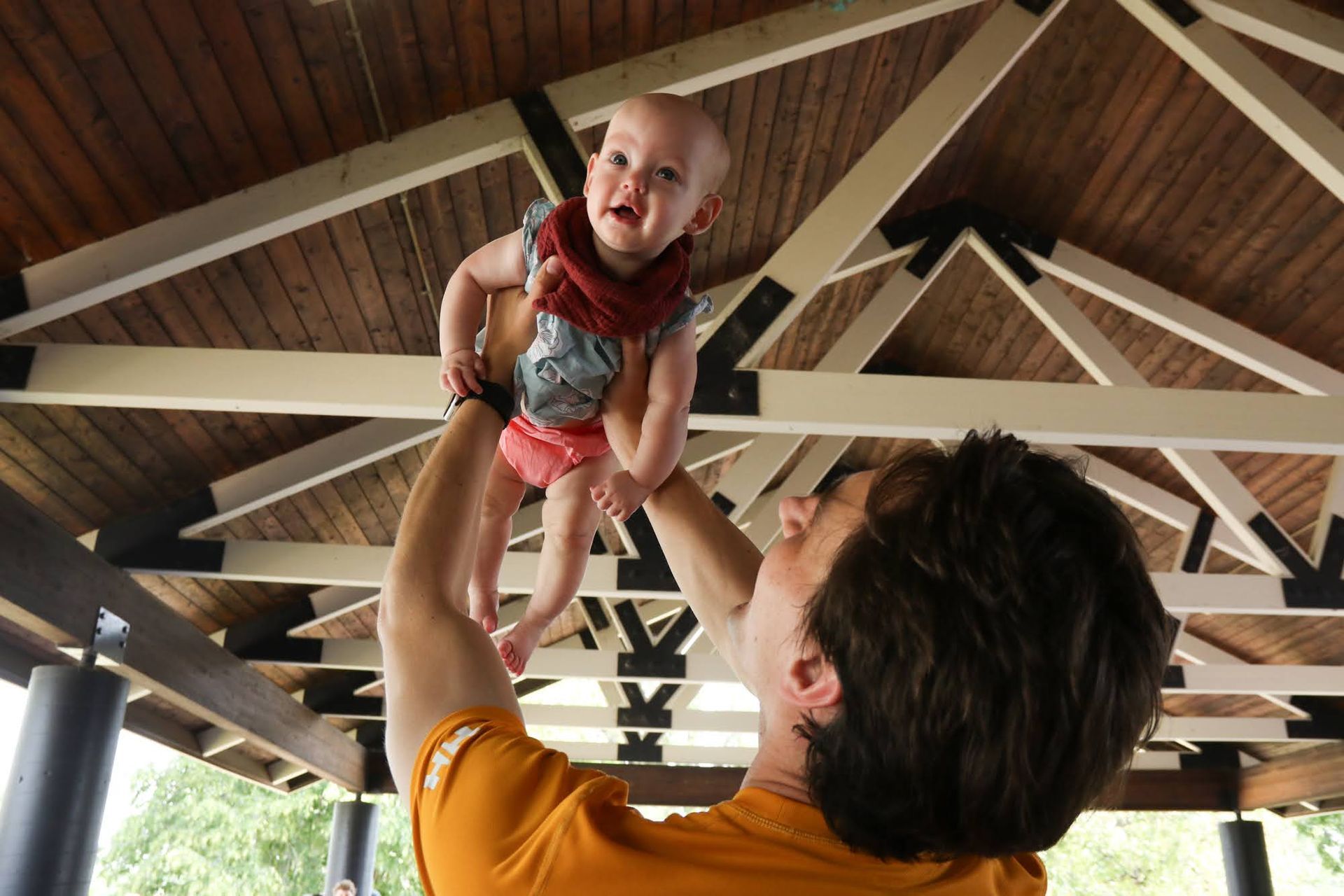 Man lifting a baby in a pavilion. Baby smiles. Wooden ceiling overhead.