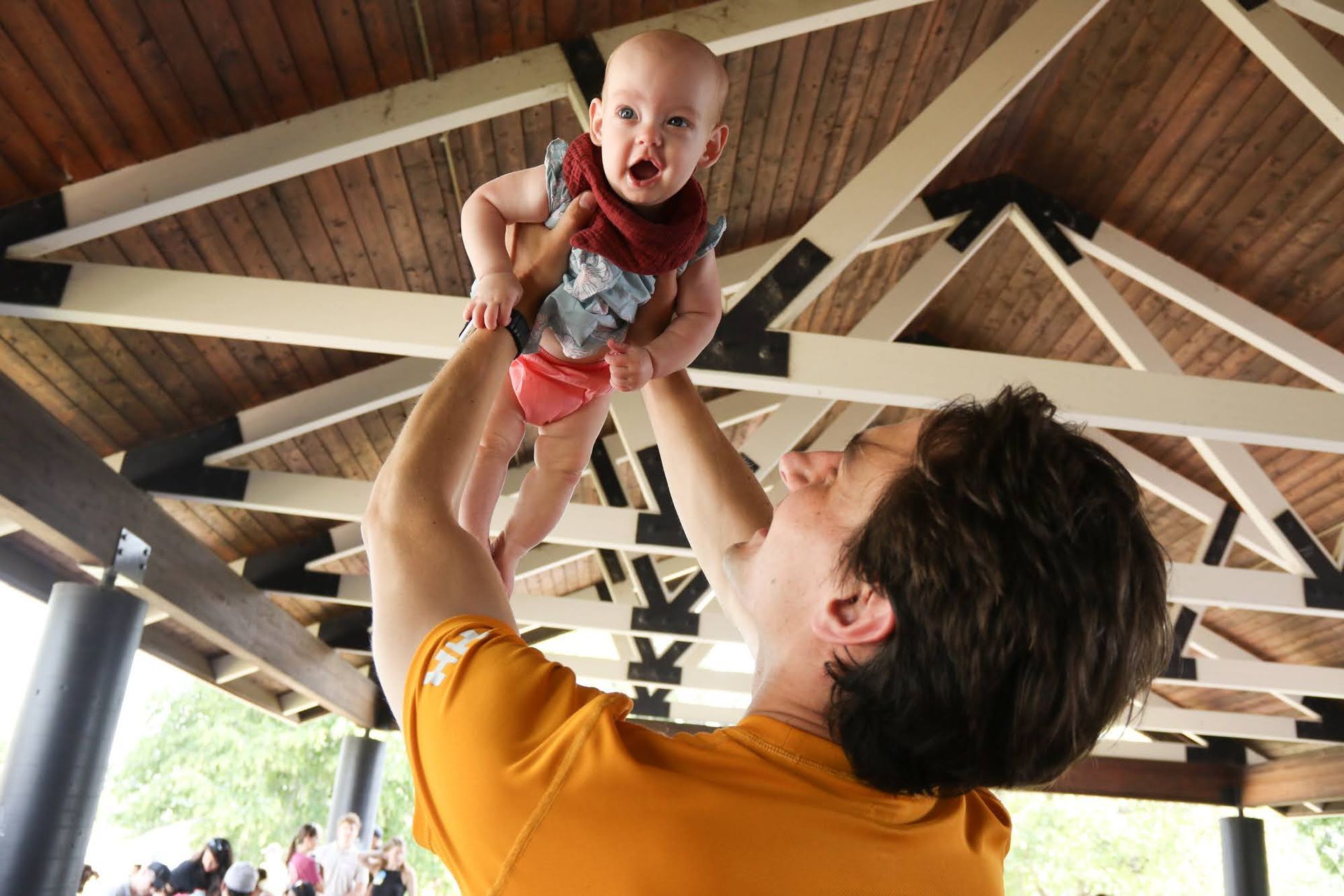 Person holding a baby up in the air under a wooden pavilion. The baby is smiling, wearing a red bib.