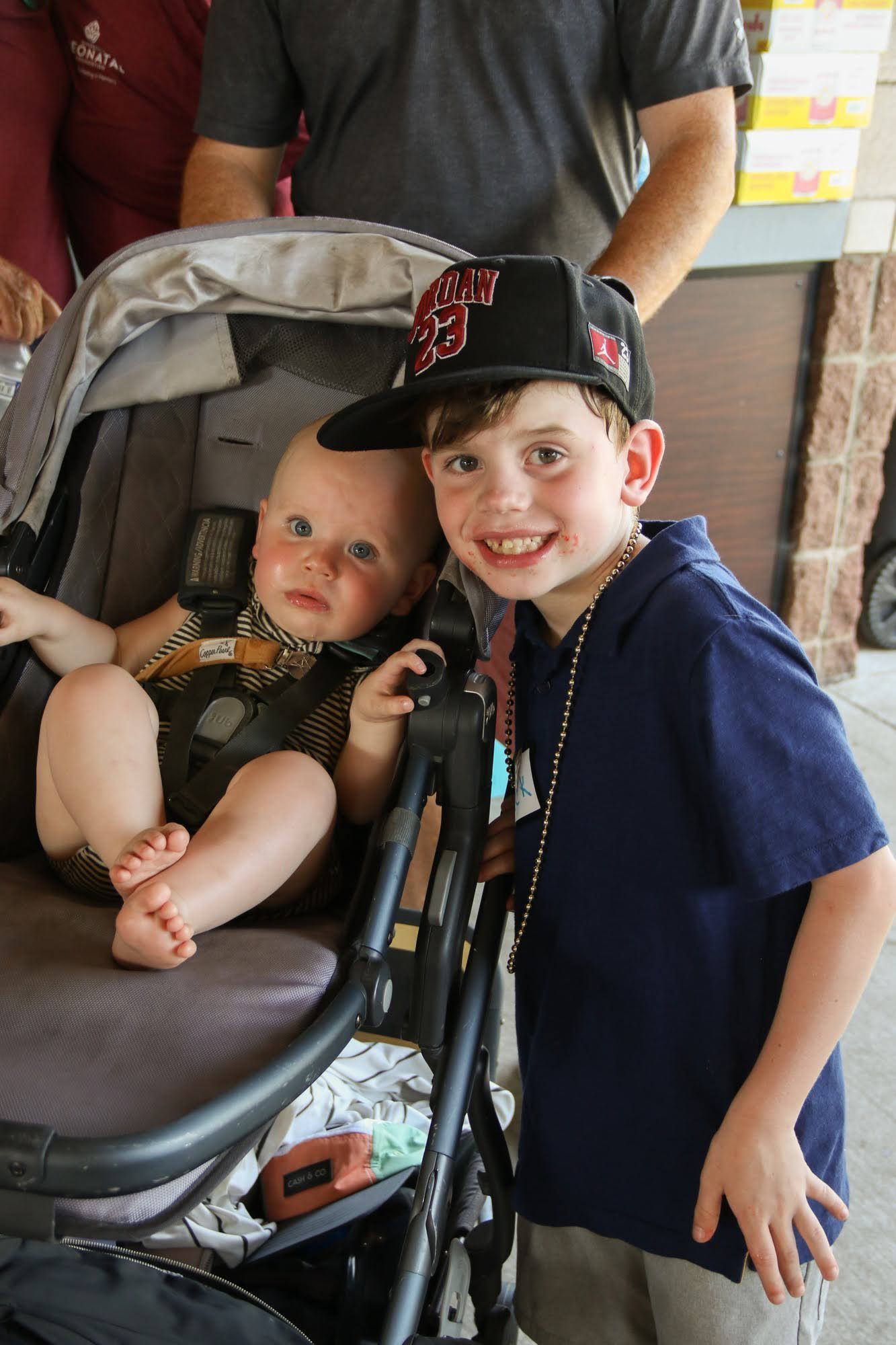 Two children: one in stroller, one smiling, wearing a baseball cap and beads. Outdoors.
