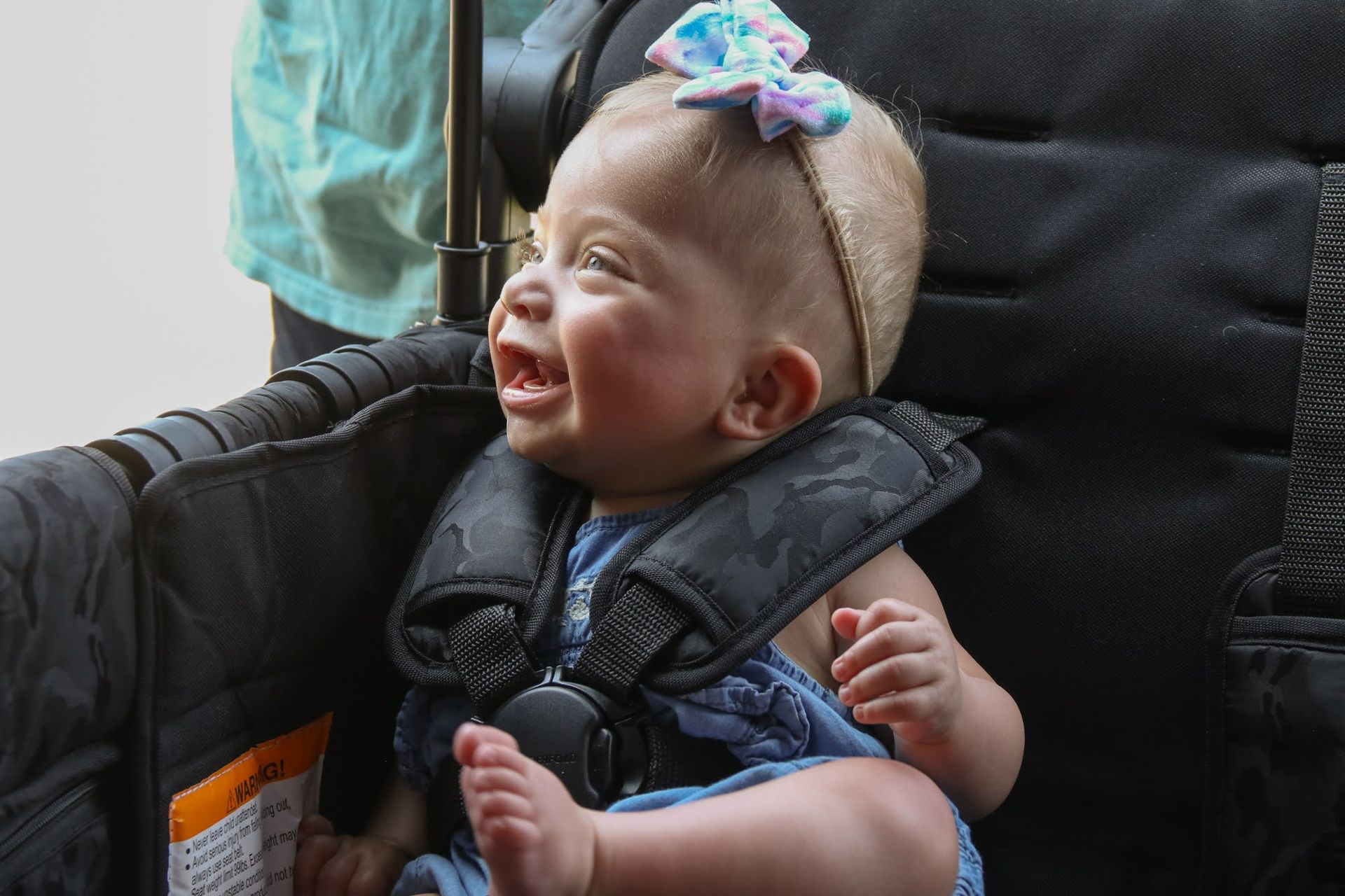 Smiling baby in a black stroller wearing a blue headband and seatbelt, looking to the side.