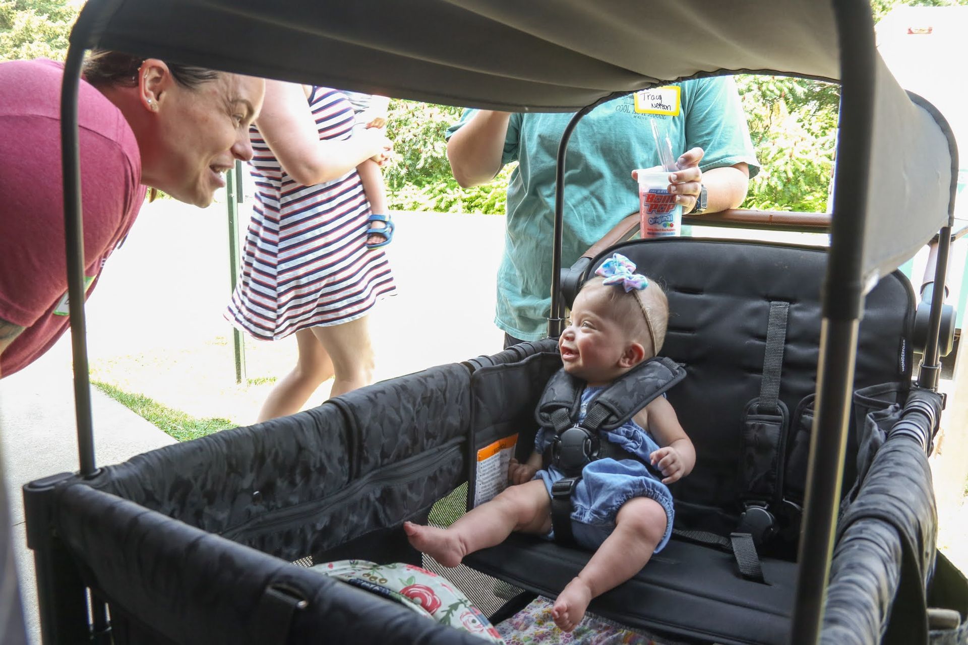 Baby in a stroller smiles as a woman leans in. Other people are in the background, outdoors.