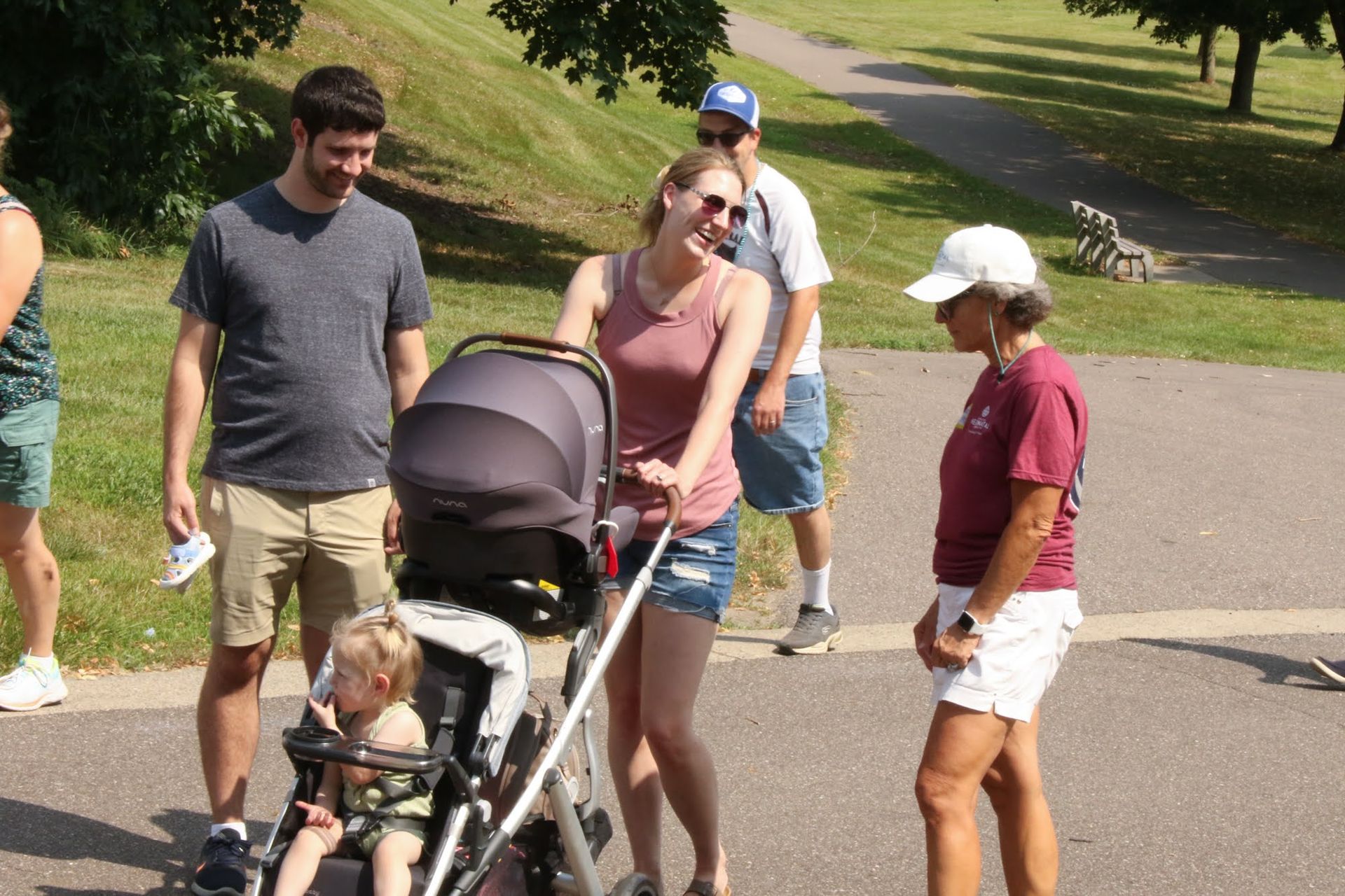 People in a park: A woman pushes a stroller, smiling. A man looks at the child, other people nearby.