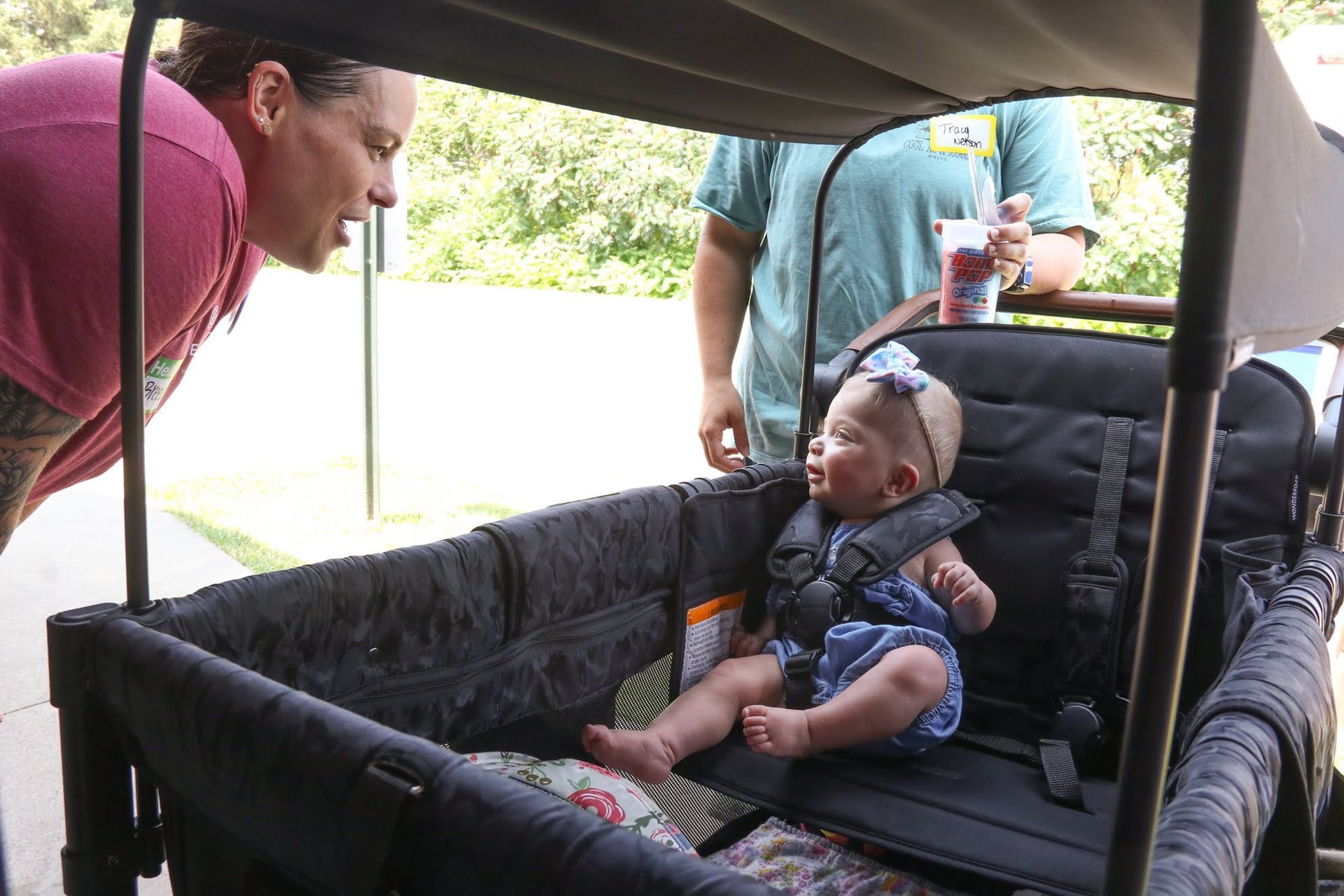 Woman leans towards baby in a stroller. Baby smiles. Another person stands nearby. Outdoors.