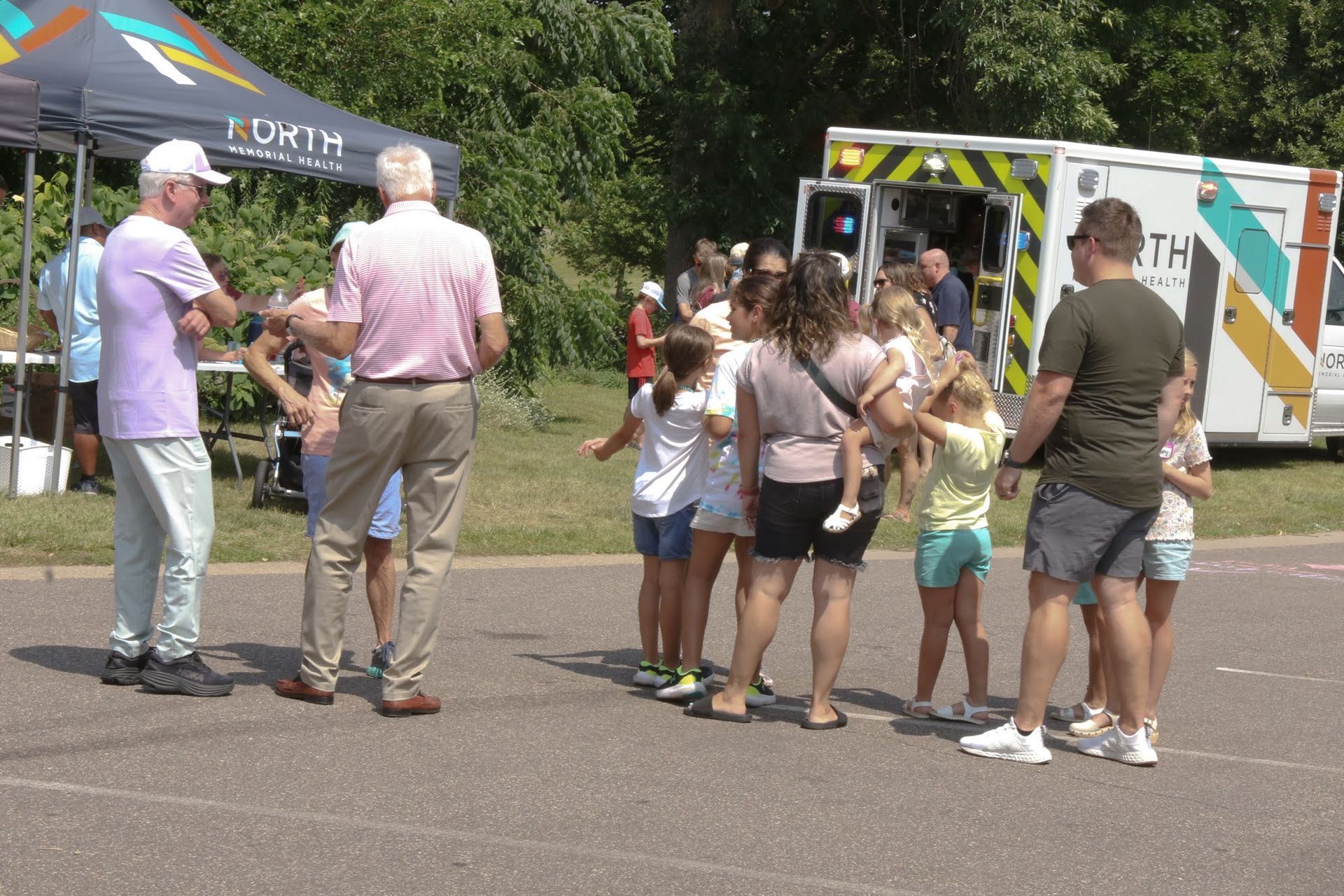 People gather near an ambulance and tent outdoors. Children interact with adults, possibly at a community event.