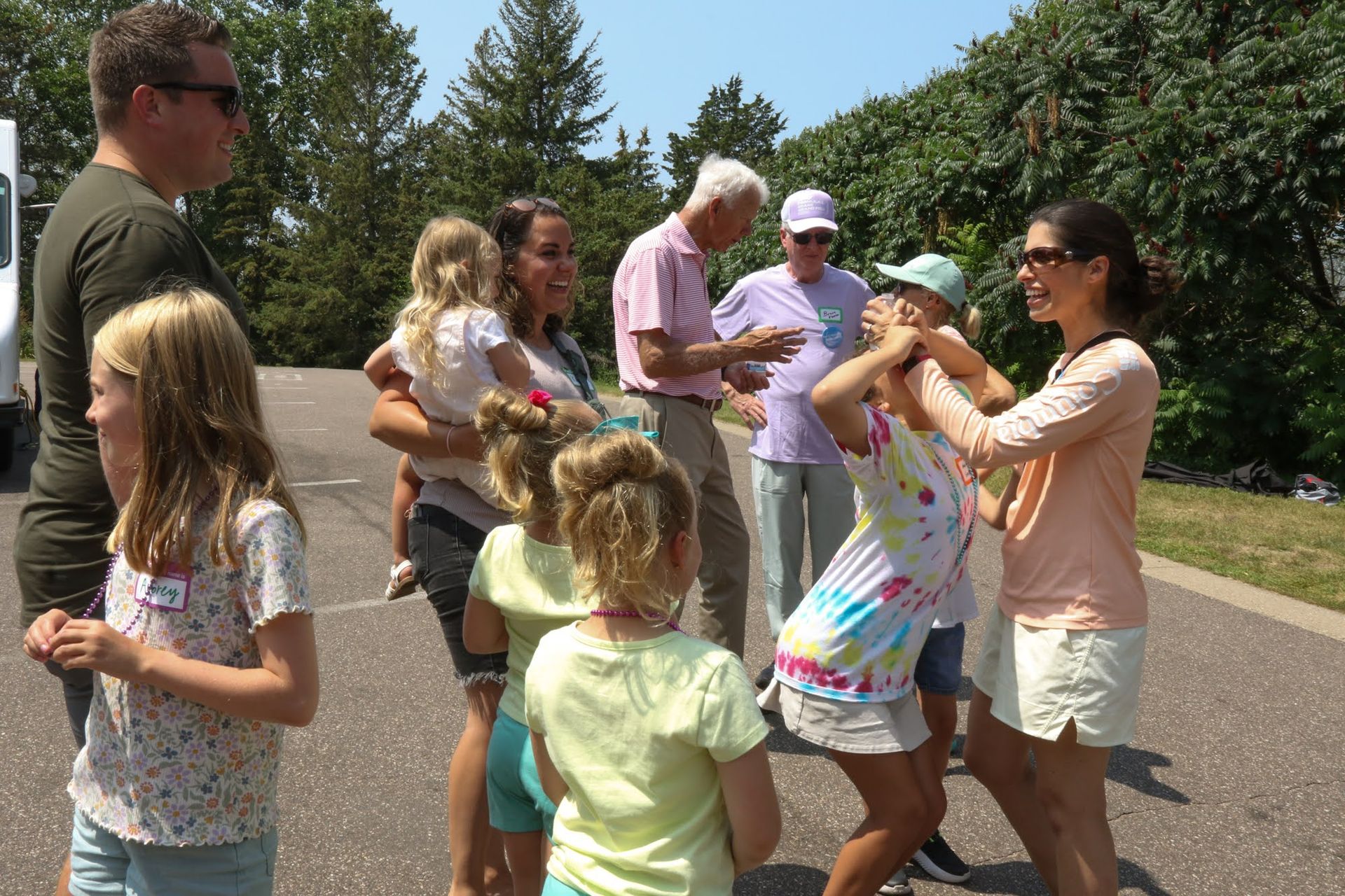 People on a paved area; two women playfully interact while others watch. Green trees and a sunny sky are in the background.