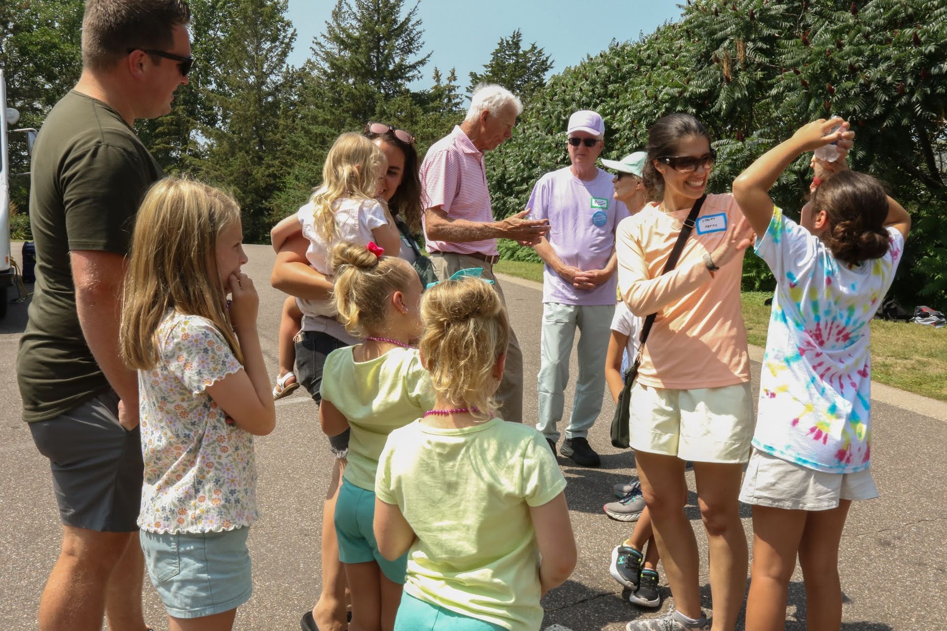 Group of people, including children, on a paved path outdoors. Two adults are interacting with the children, one holding a string with something on it.