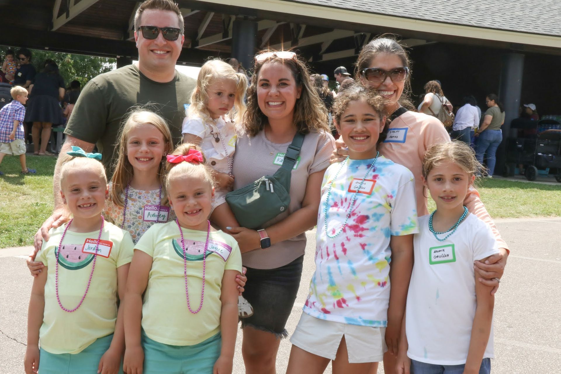 Group of people posing outdoors; smiling, wearing casual clothes. Some children in front, adults behind.